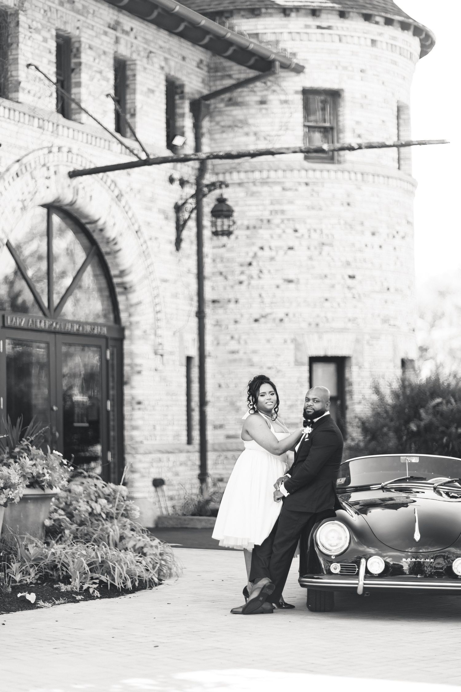 Black and white photo of bride and groom posing beside vintage Porsche at brick venue.