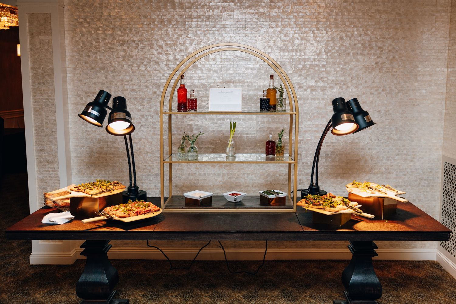 Wedding reception buffet table with food warmers and lamps against a shimmering mosaic wall.