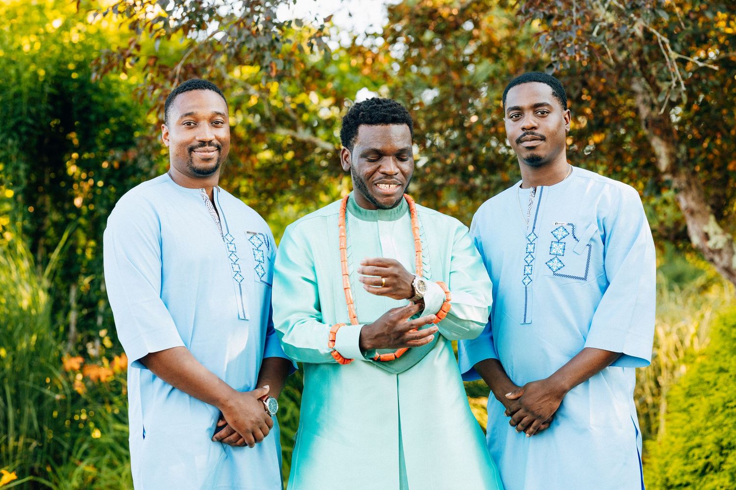 Three men in light blue traditional African attire smile outdoors, with the center man wearing a mint green outfit.