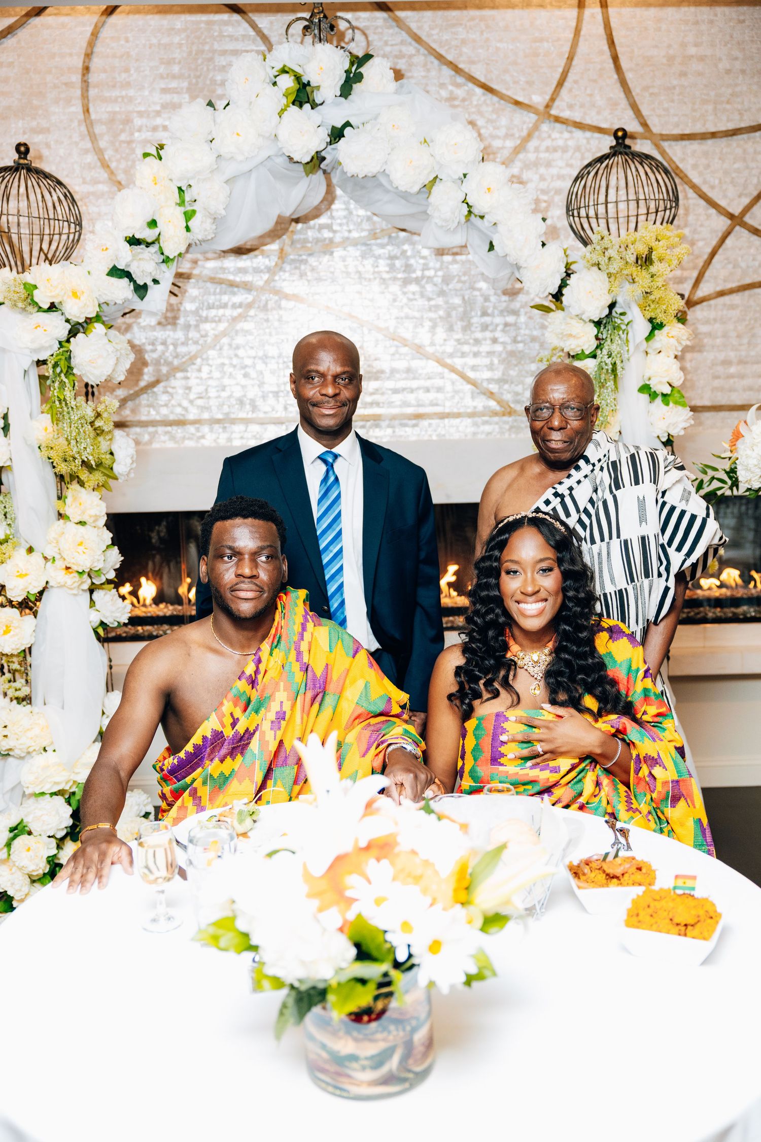Bride and groom in Kente attire pose with two elder men in suits at a traditional African wedding reception.