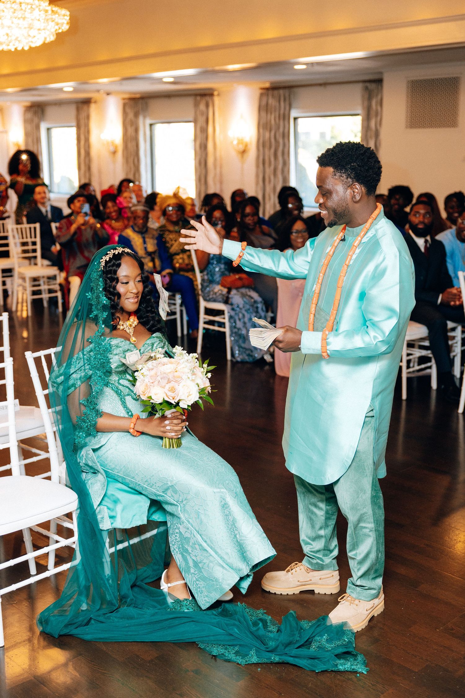 A groom in a teal African suit gestures to his seated bride in a matching teal gown and veil.