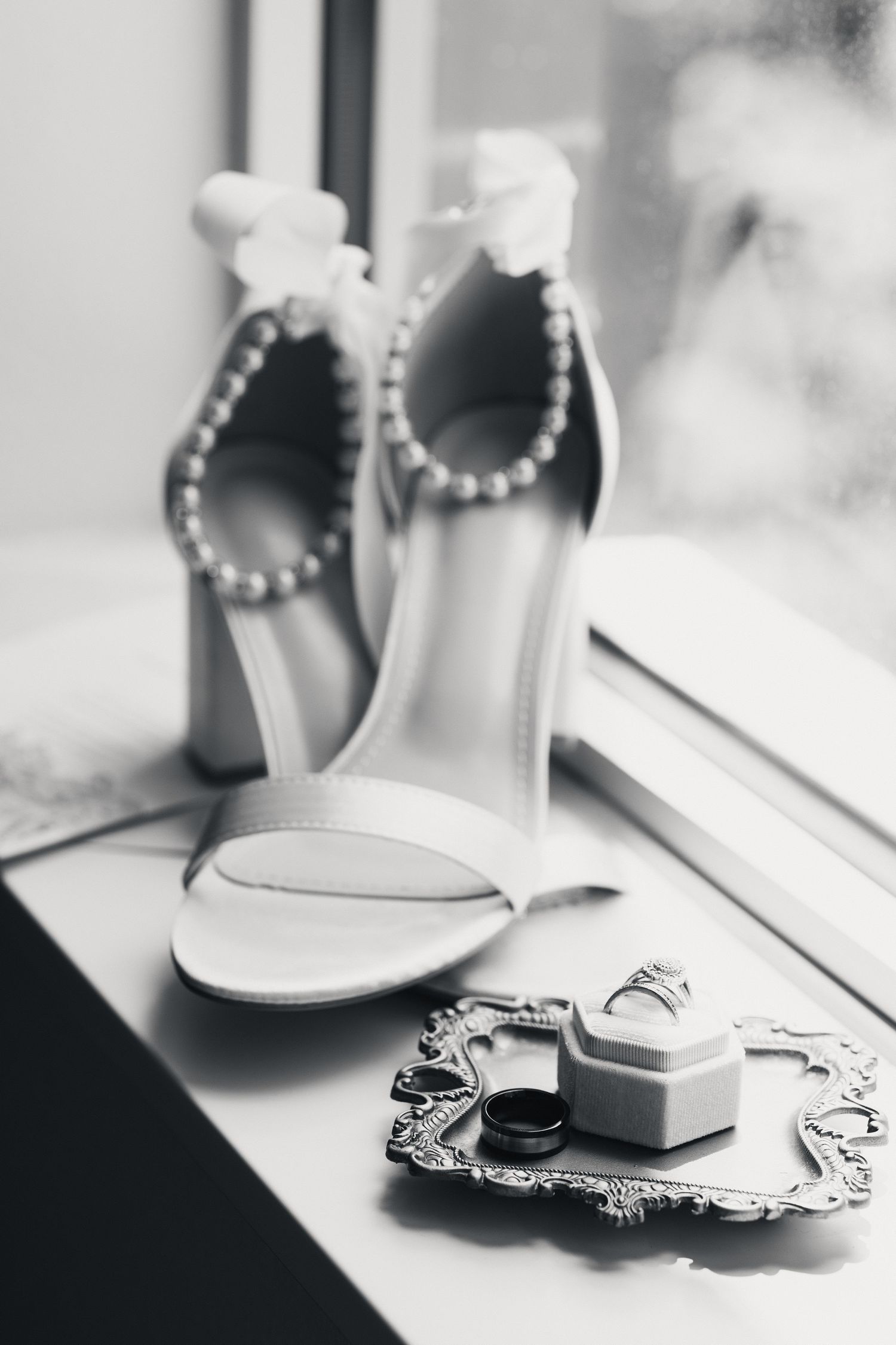 Black and white photo of bridal heels with pearl ankle straps beside wedding rings on a decorative tray by a window.