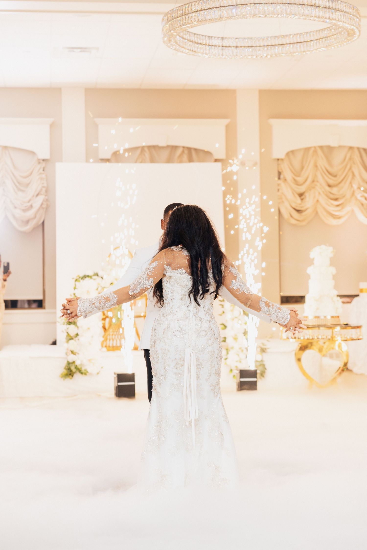 Bride in lace wedding gown stands on foggy dance floor with arms outstretched at elegant white and gold reception.