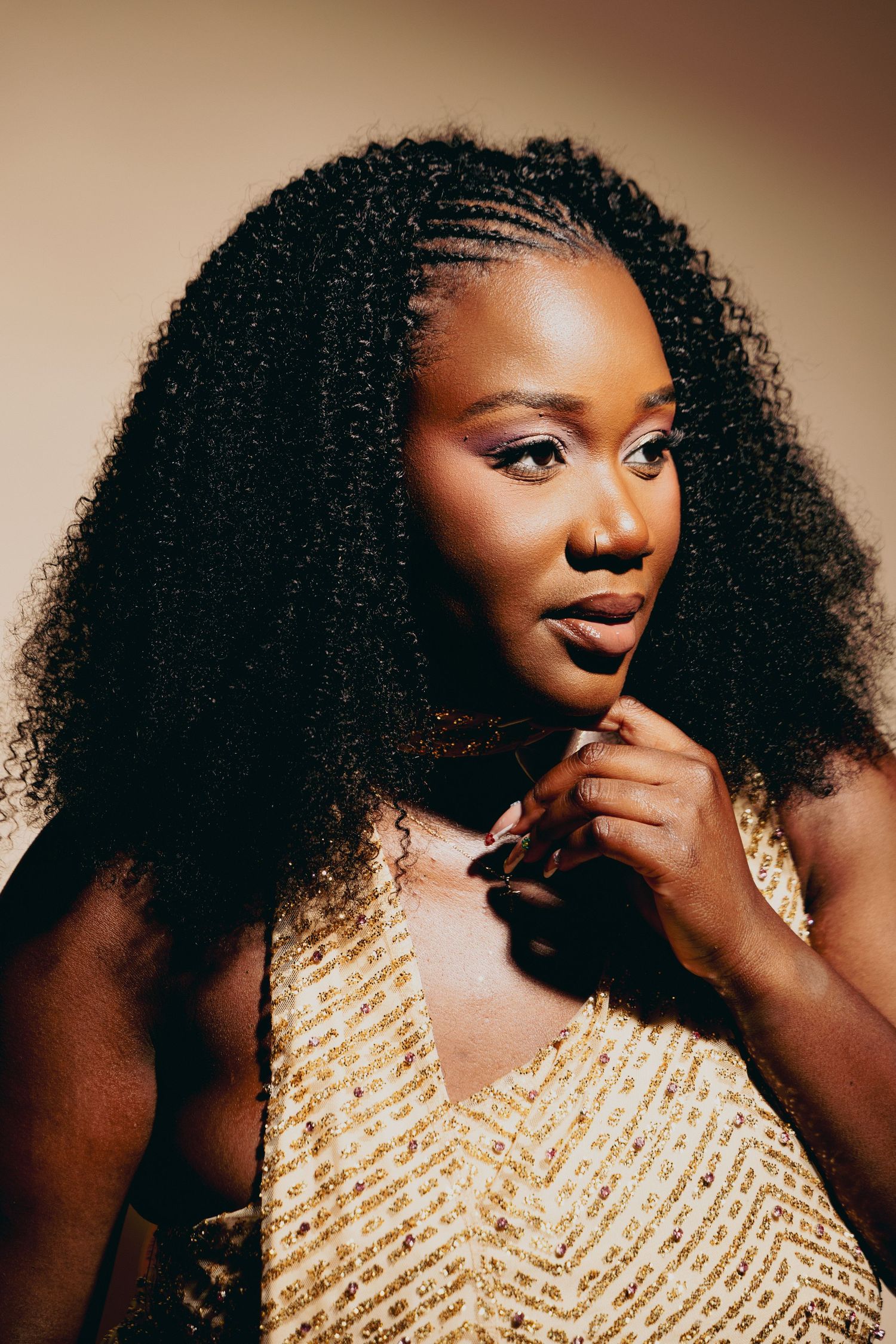 A young Black woman with curly braided hair poses confidently in a gold sequined top against a warm beige background.