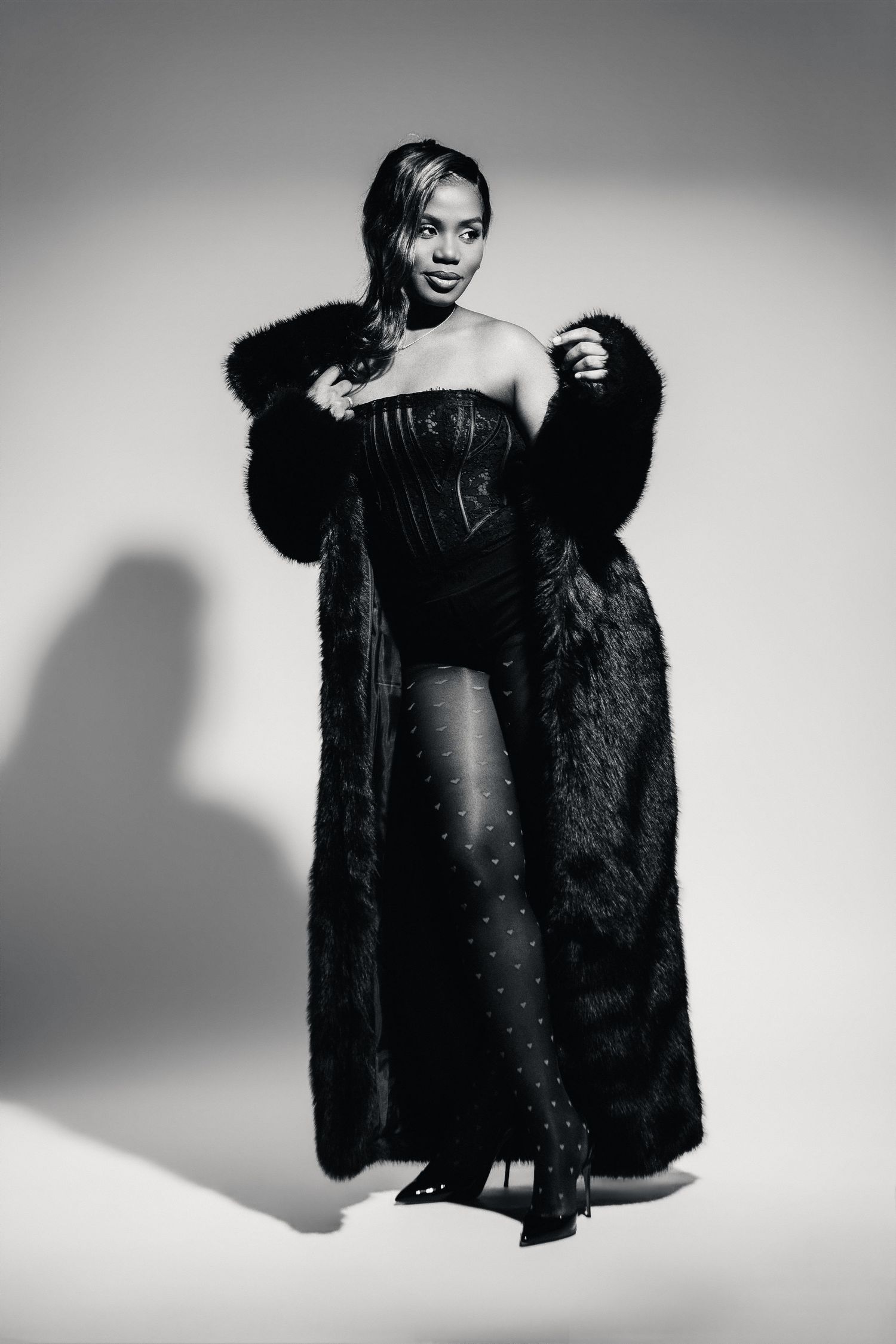 Black and white photo of a woman in a strapless corset and long fur coat posing confidently in a studio.