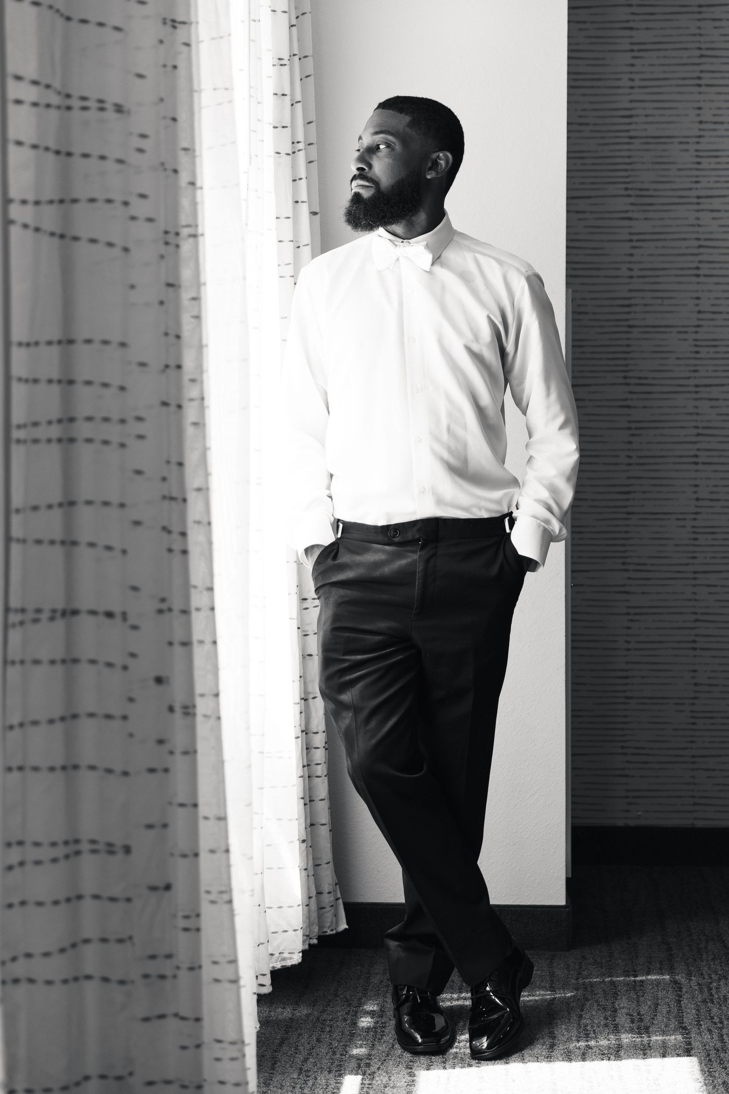 Black and white portrait of a bearded man in white dress shirt and black trousers standing by a curtained window.
