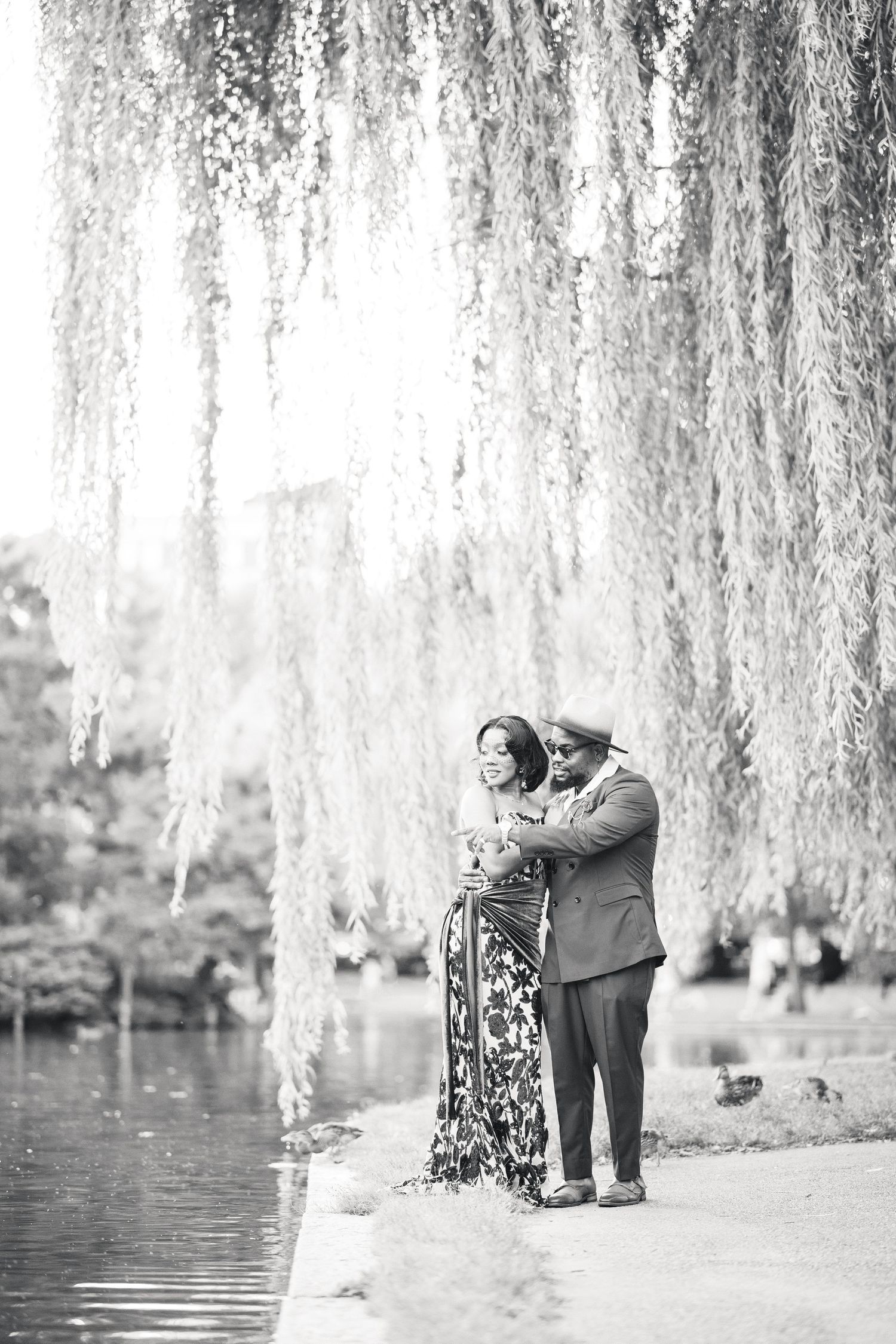 Black and white photo of couple embracing tenderly under a weeping willow tree beside a tranquil lake.