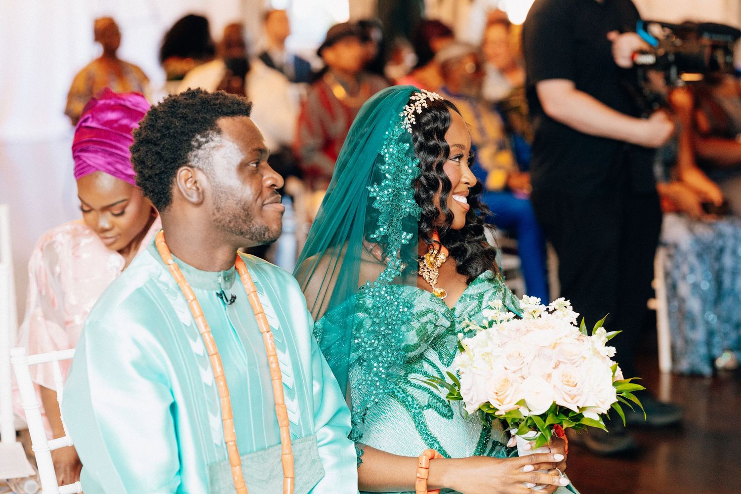 Couple in matching teal traditional African wedding attire seated together, bride holding white and pink bouquet.