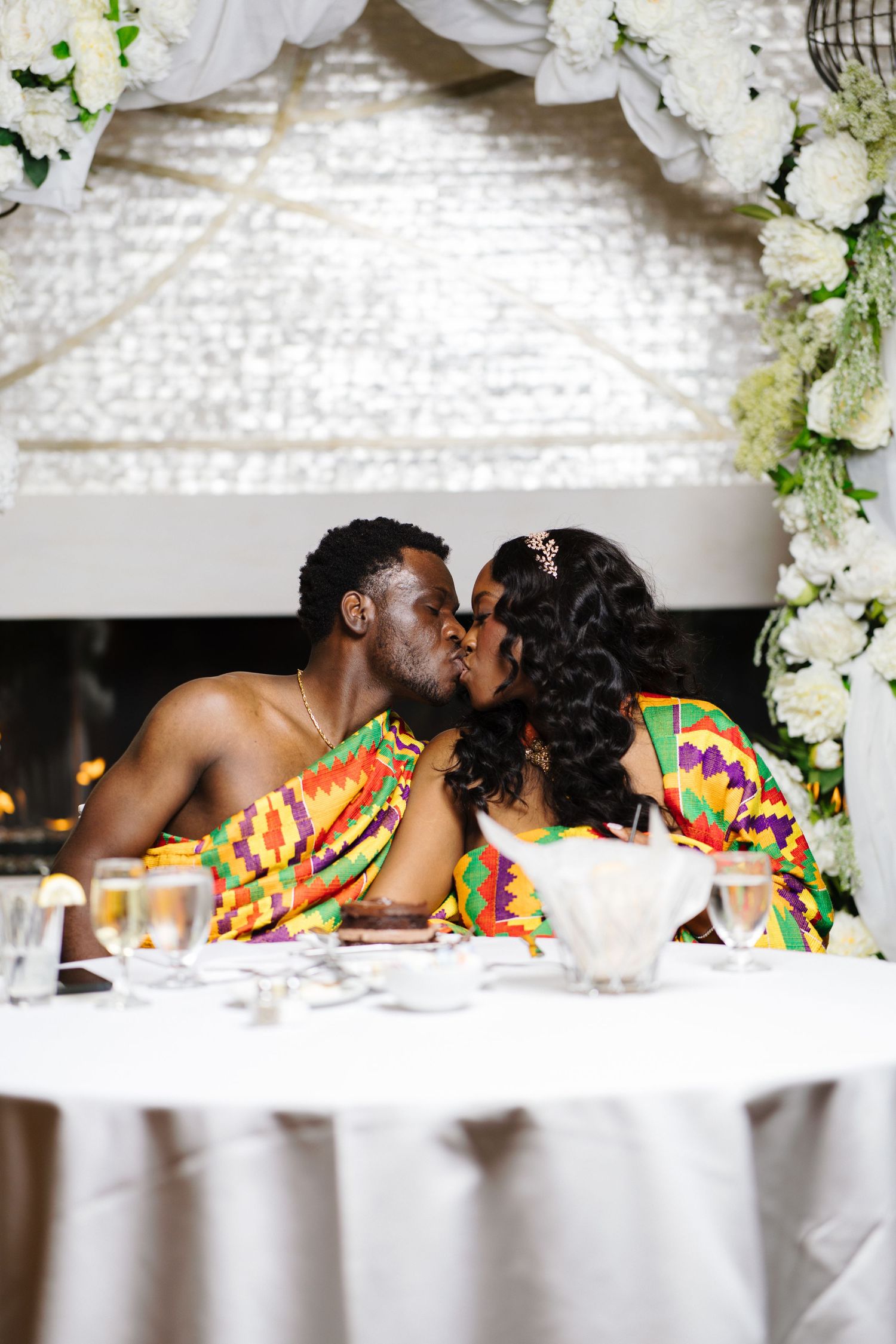 Newlywed couple sharing a kiss at their sweetheart table adorned with white floral arch decor.