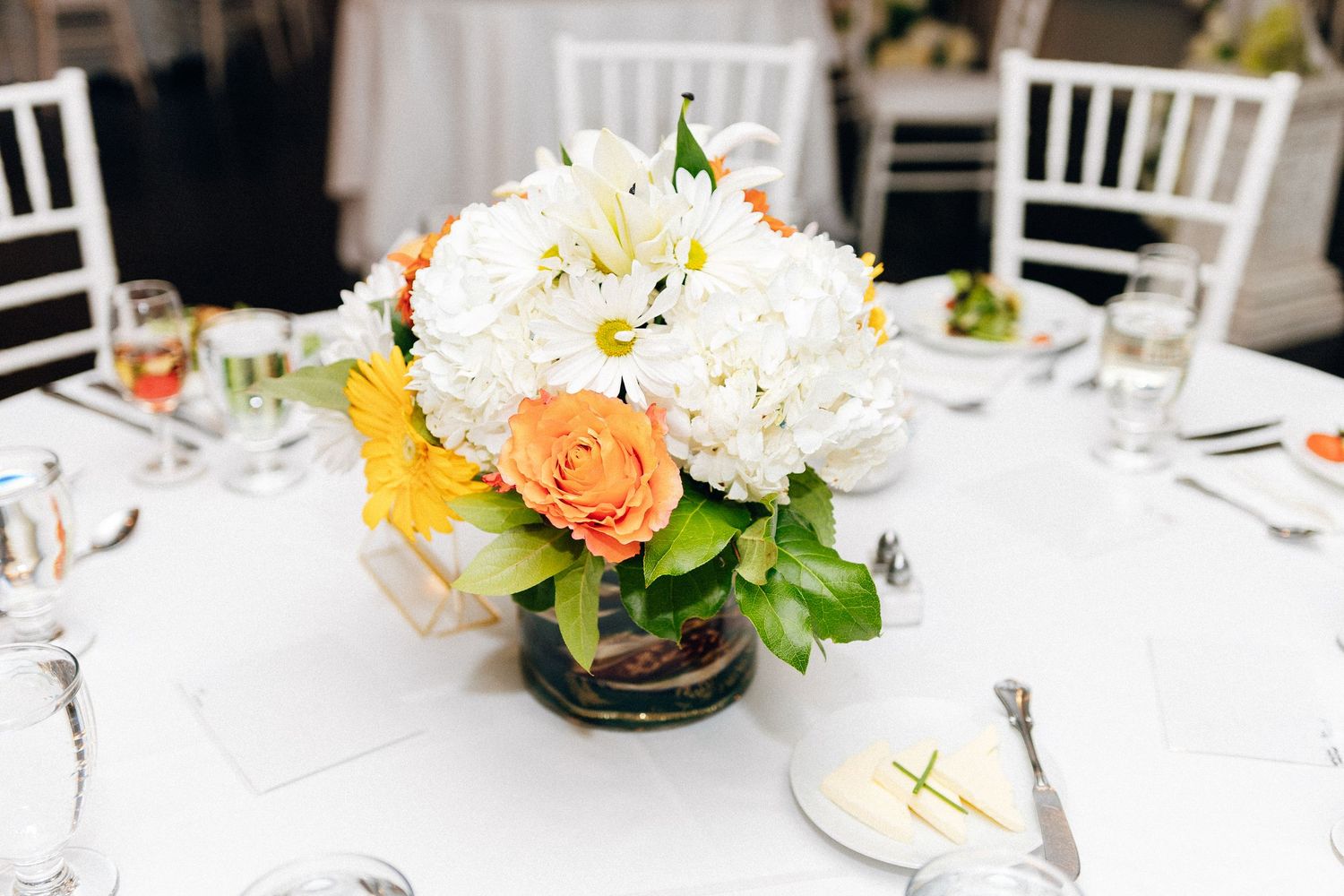 Elegant wedding reception centerpiece with white hydrangeas, orange roses, and green leaves in a glass vase on a white tablecloth.