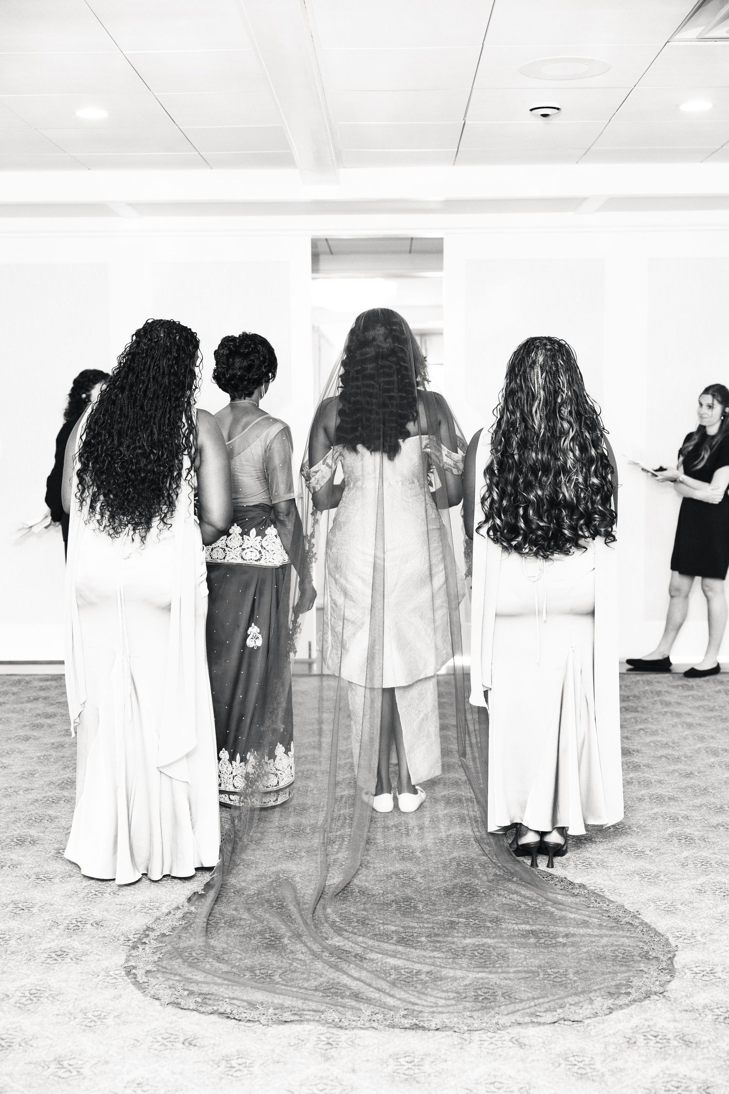 Bridesmaids stand behind a bride in a flowing lace gown and veil in a black and white wedding photo.