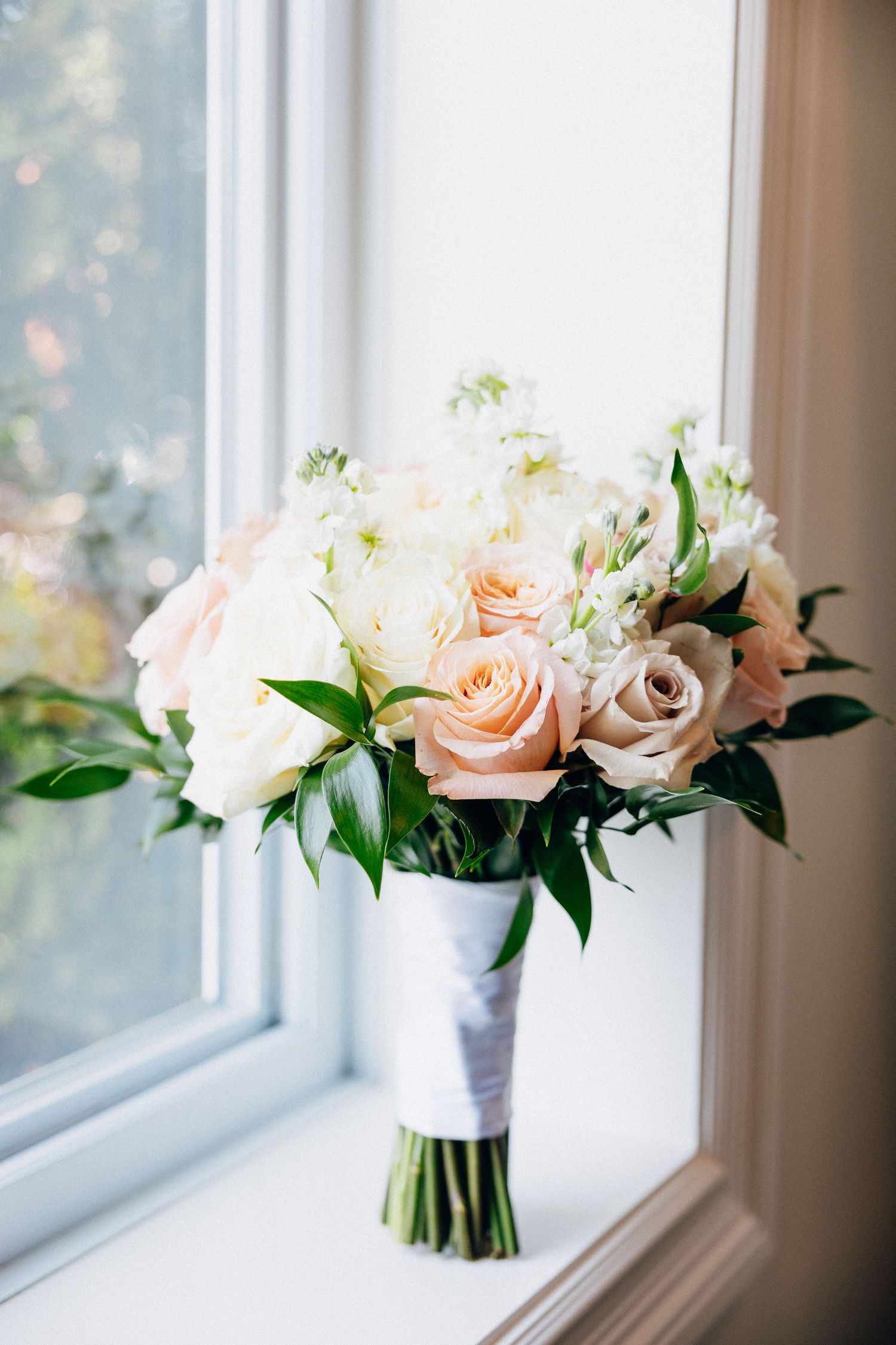 Elegant wedding bouquet of peach and white roses with greenery, resting on a sunlit window sill.