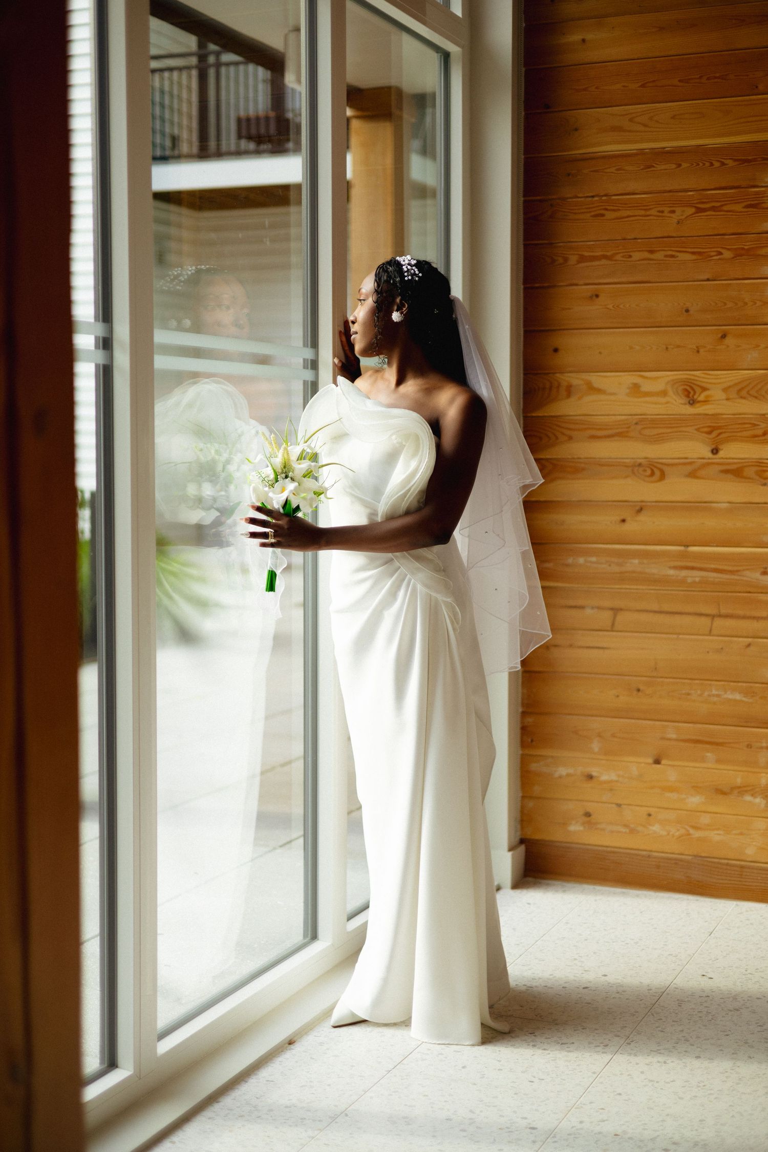 Stunning Black bride in white wedding dress and veil gazes out large window holding floral bouquet.
