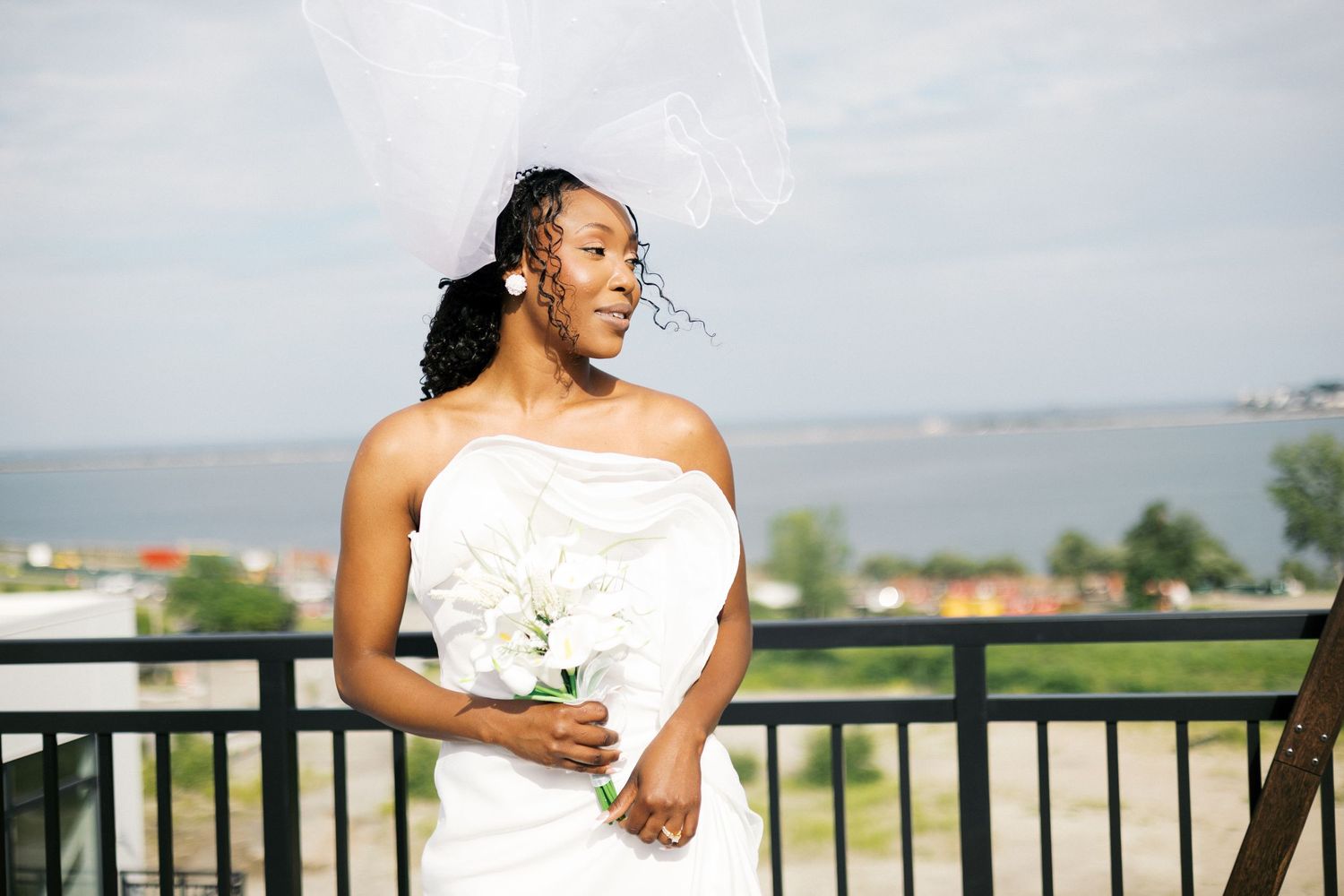 Bride in strapless white gown holds bouquet on balcony overlooking water, veil blowing in wind.
