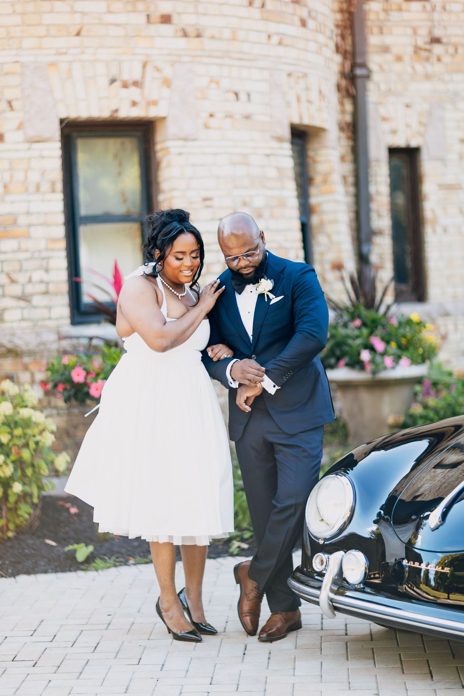 Black bride and groom in wedding attire share a tender moment beside a vintage classic car outside a stone building.
