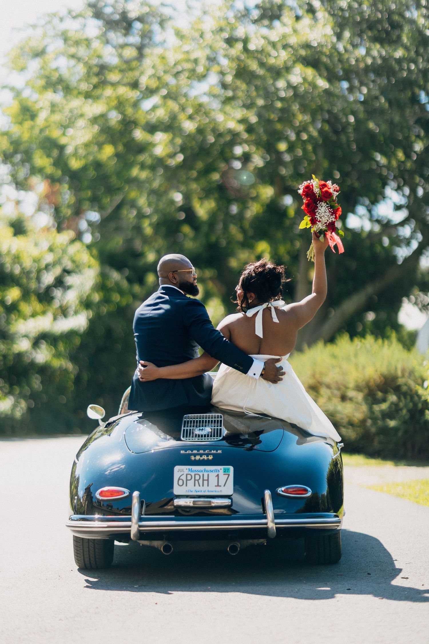 Newlyweds celebrate atop a classic blue Porsche convertible as bride raises red bouquet in joyful triumph.