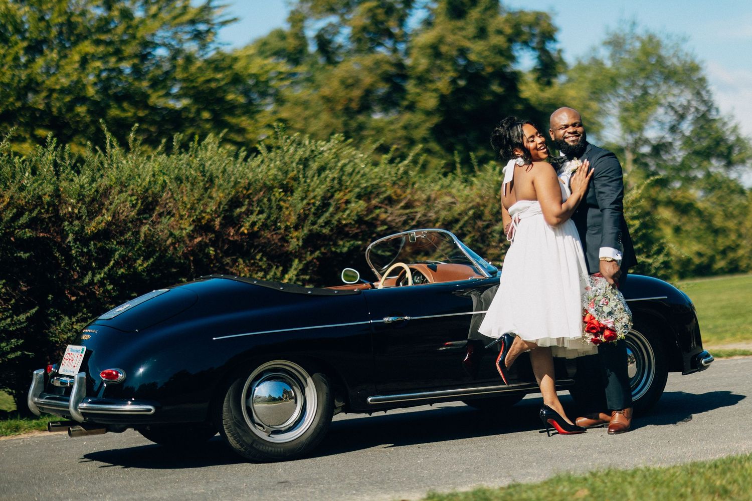 Happy bride and groom embracing beside a vintage black Porsche 356 convertible on a sunny day.