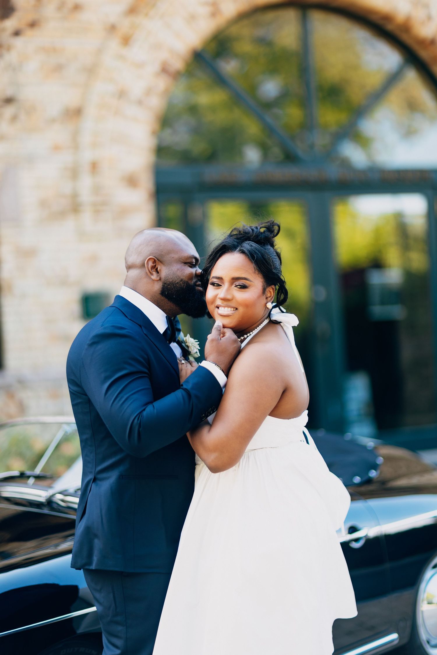 Smiling bride in white halter gown poses with groom whispering in her ear by a vintage car.