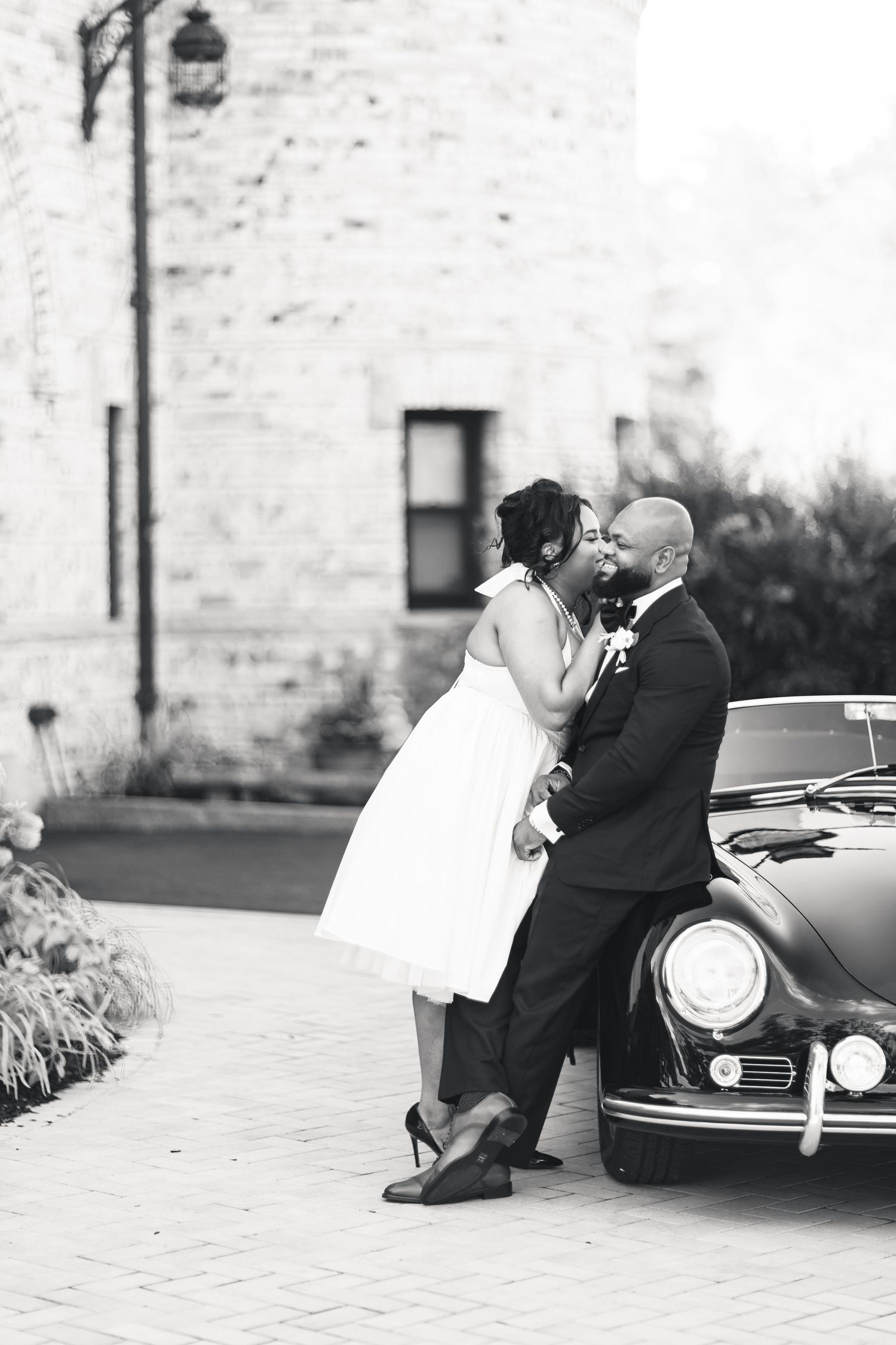 Black and white photo of bride and groom kissing beside a classic vintage car at stone venue.