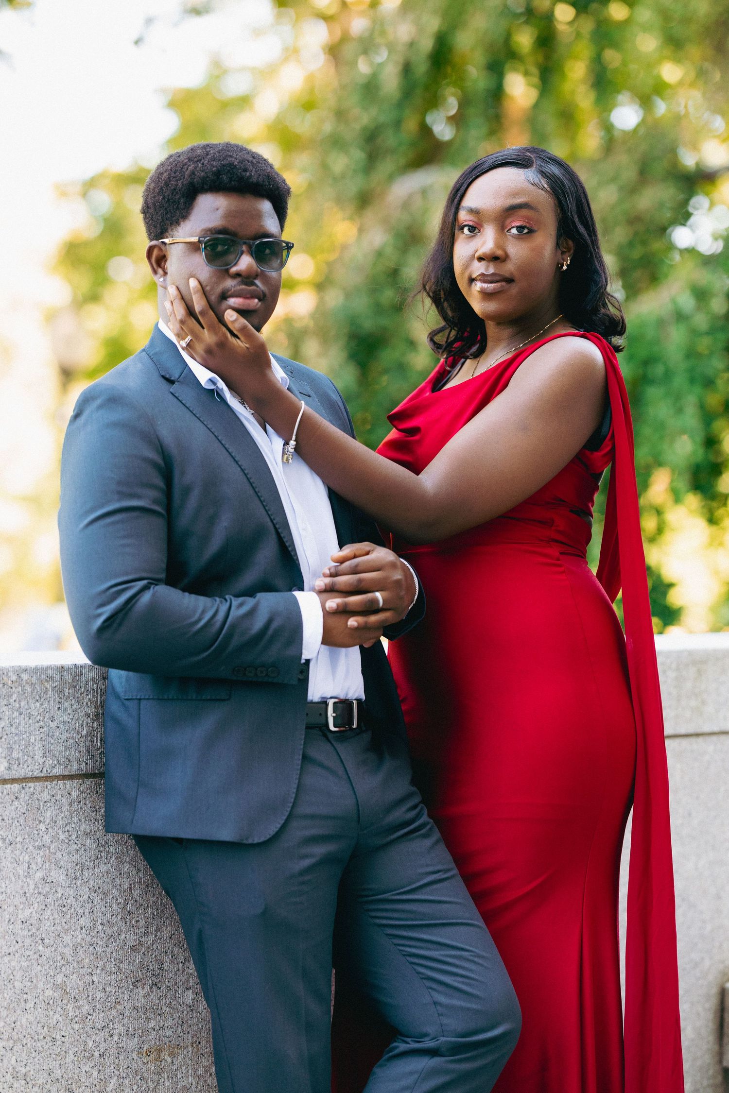 Elegant Black couple posing outdoors, man in gray suit with sunglasses, woman in red gown, green trees background.