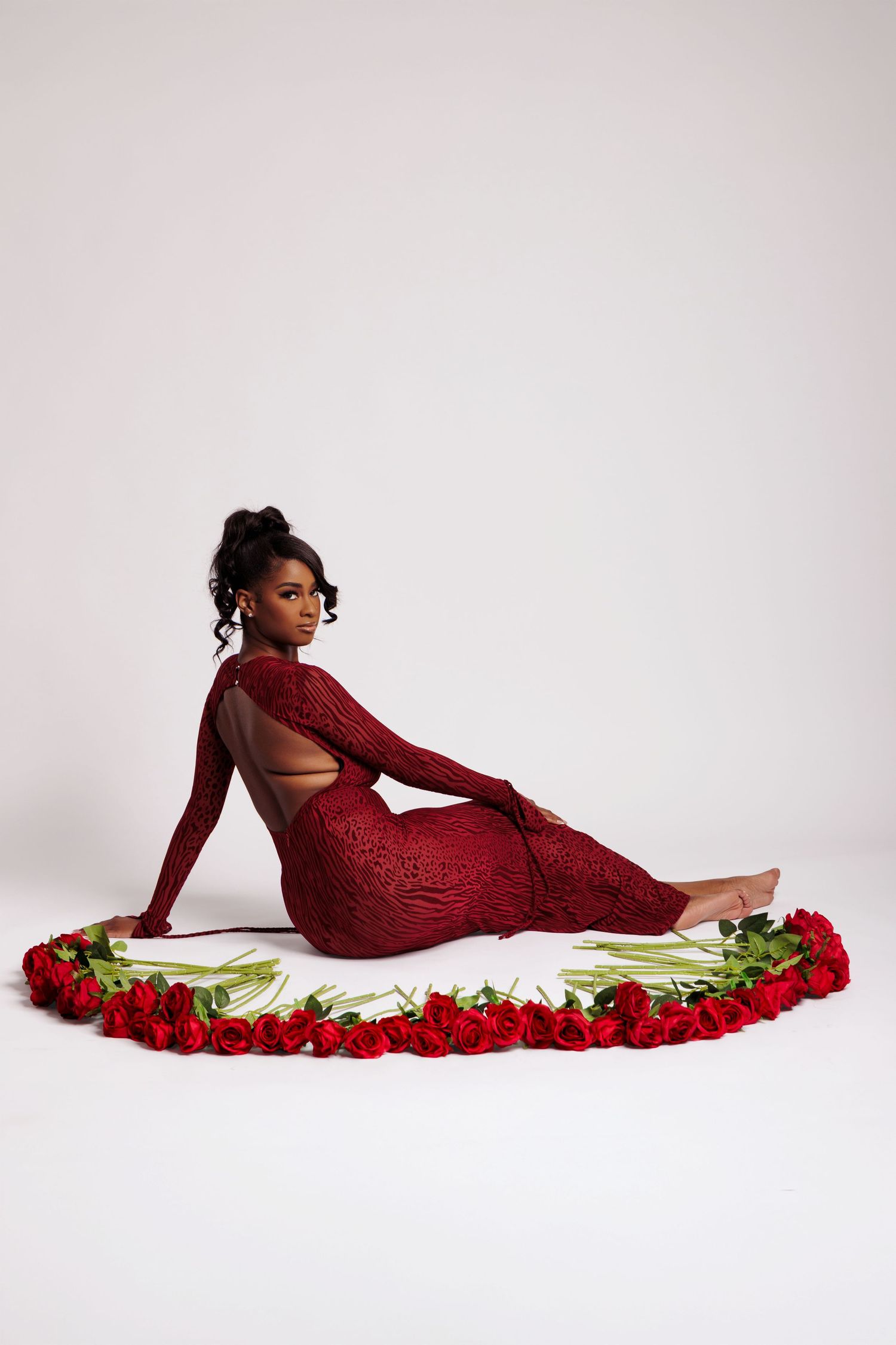 Woman in red sequin gown poses on floor surrounded by red roses against white background.