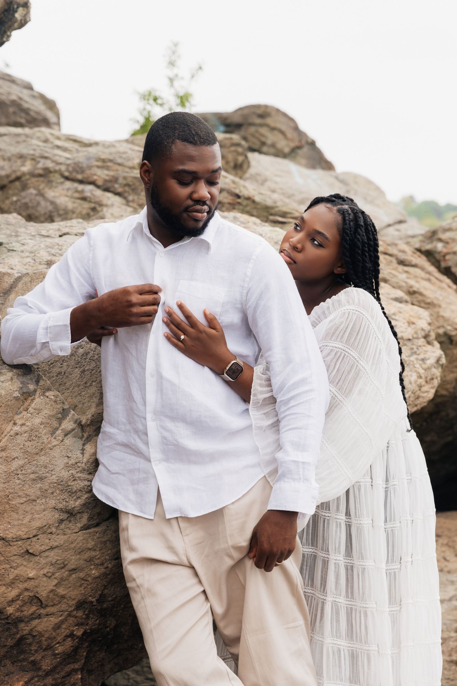 A Black couple in matching white outfits poses romantically against large rocks outdoors.