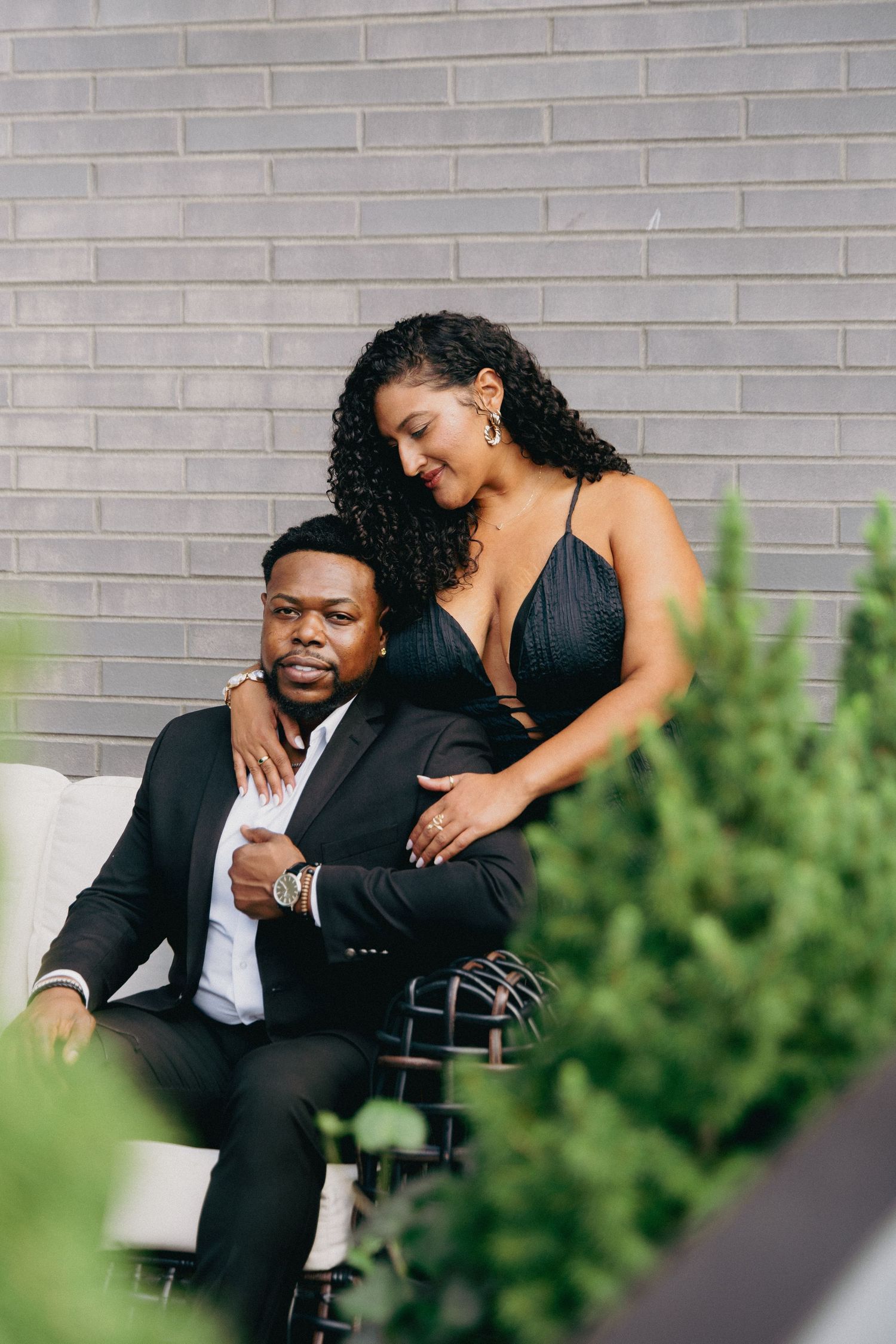 Stylish couple in black formal wear sharing an intimate moment on a wicker sofa with lush greenery nearby.