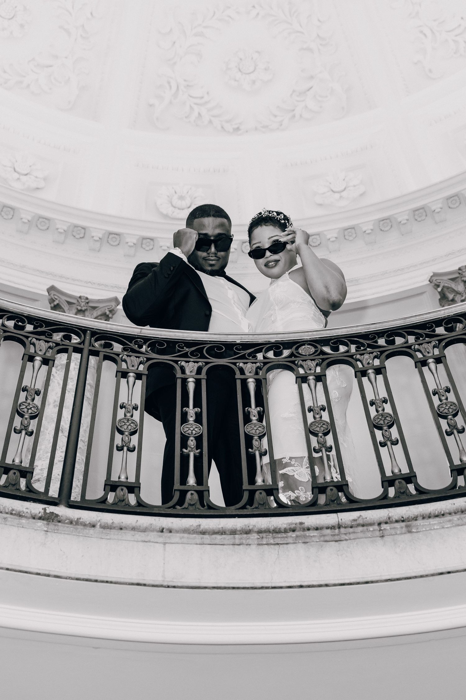 Black and white photo of bride and groom wearing sunglasses on ornate balcony in domed building.