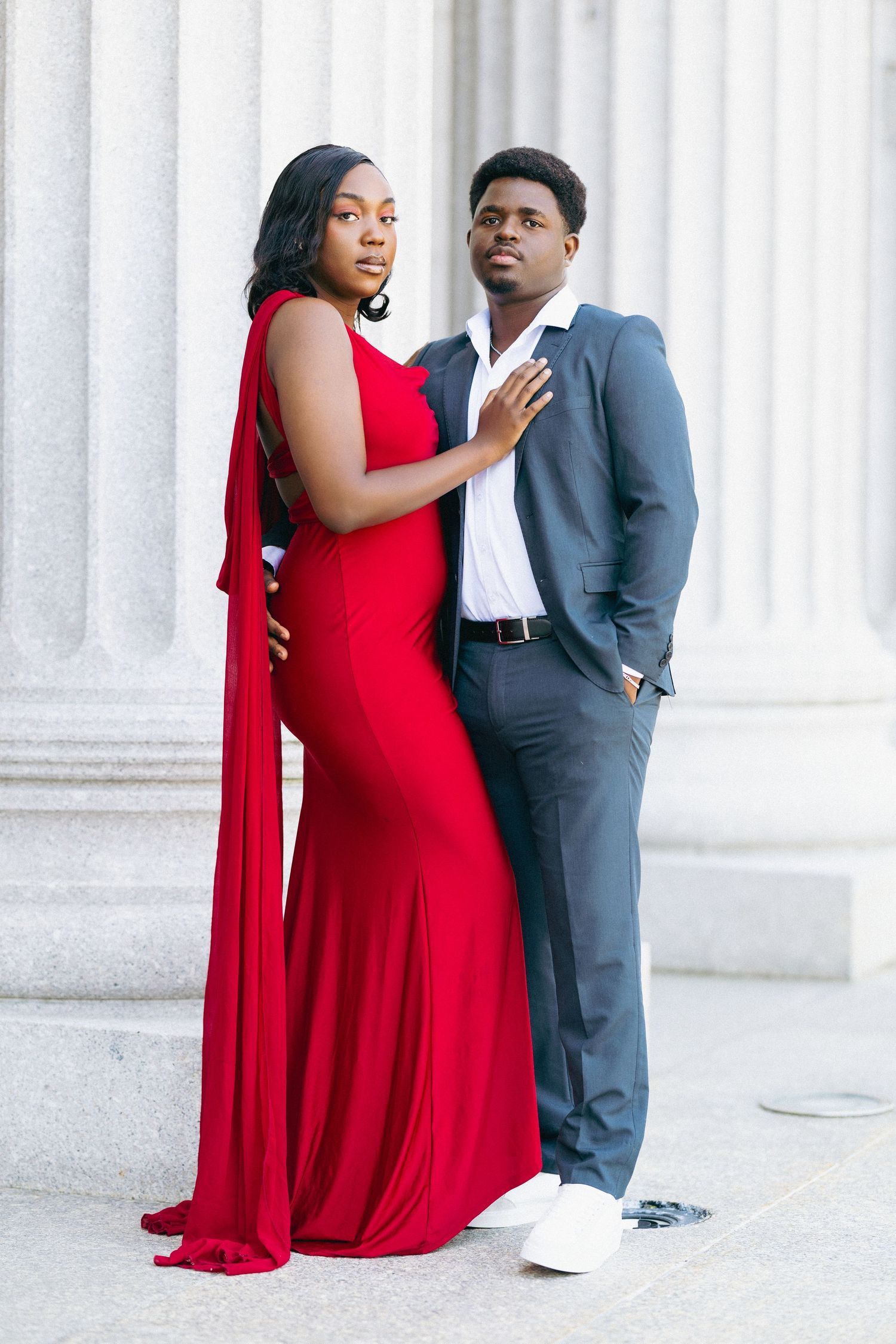 Elegant couple posing by columns, woman in red gown, man in gray suit, formal portrait photography.