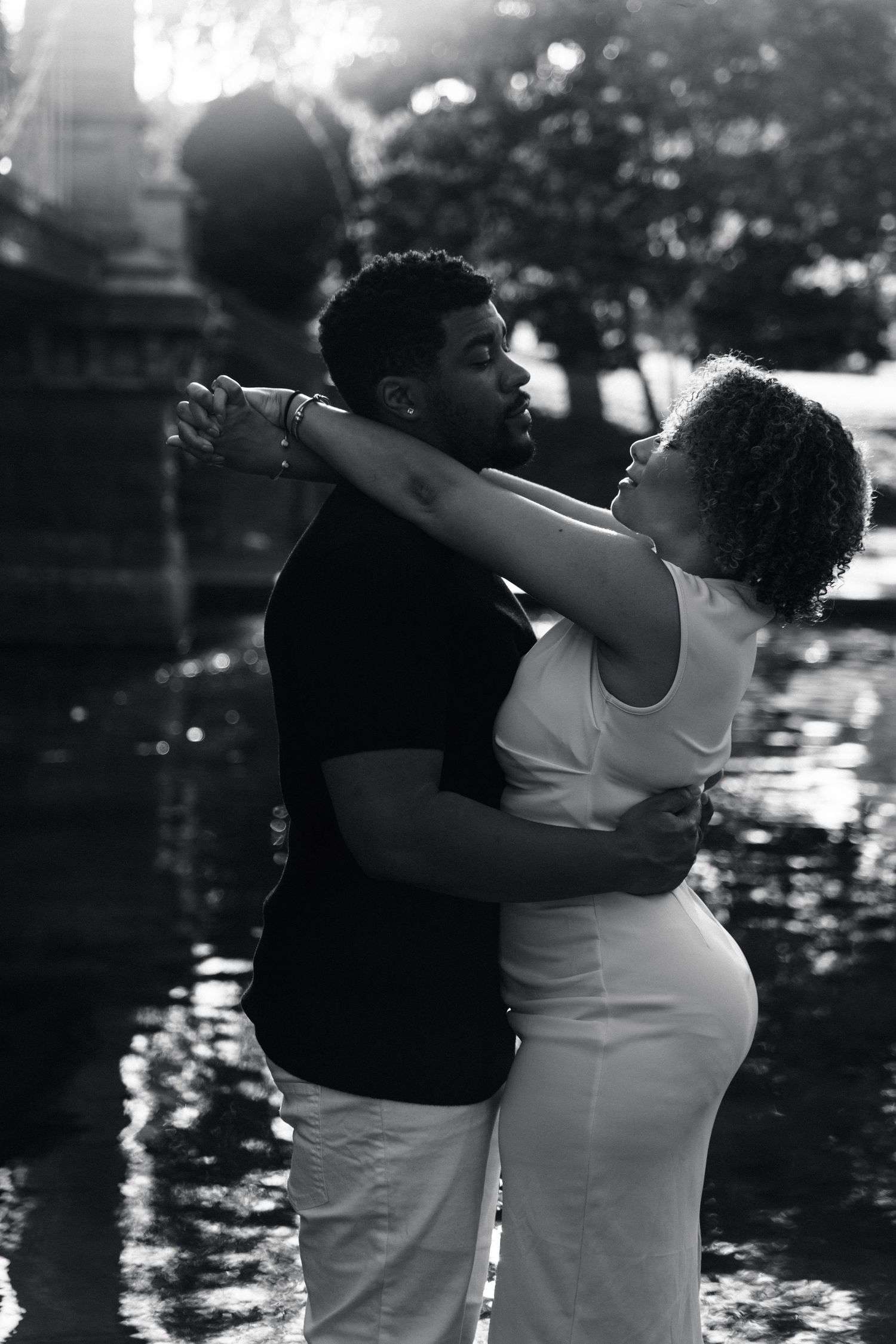 Black and white photo of a couple embracing near a fountain in a sunlit park setting.