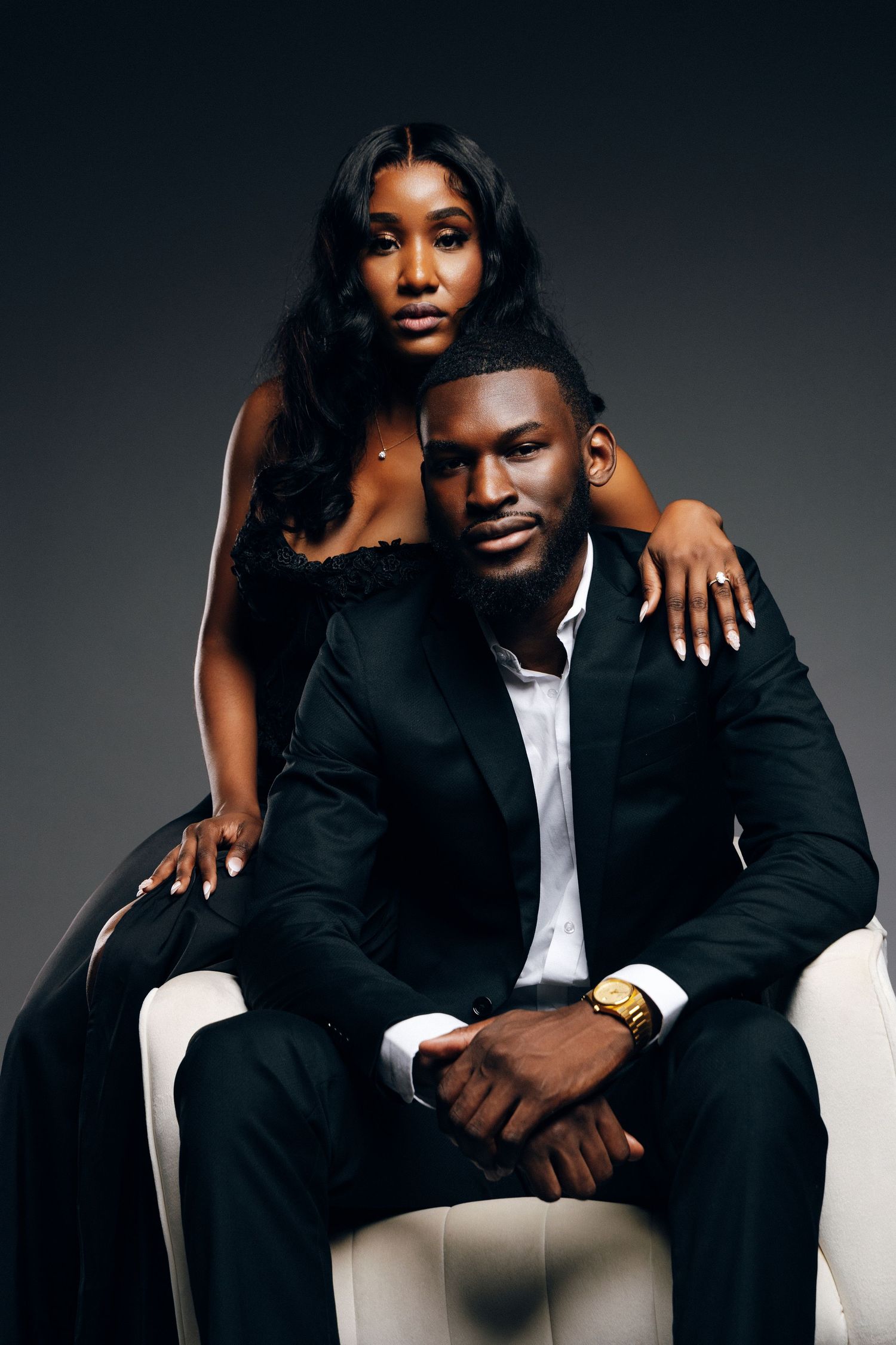 Elegant Black couple in formal attire, man seated in white chair wearing suit and gold watch, woman standing behind him.