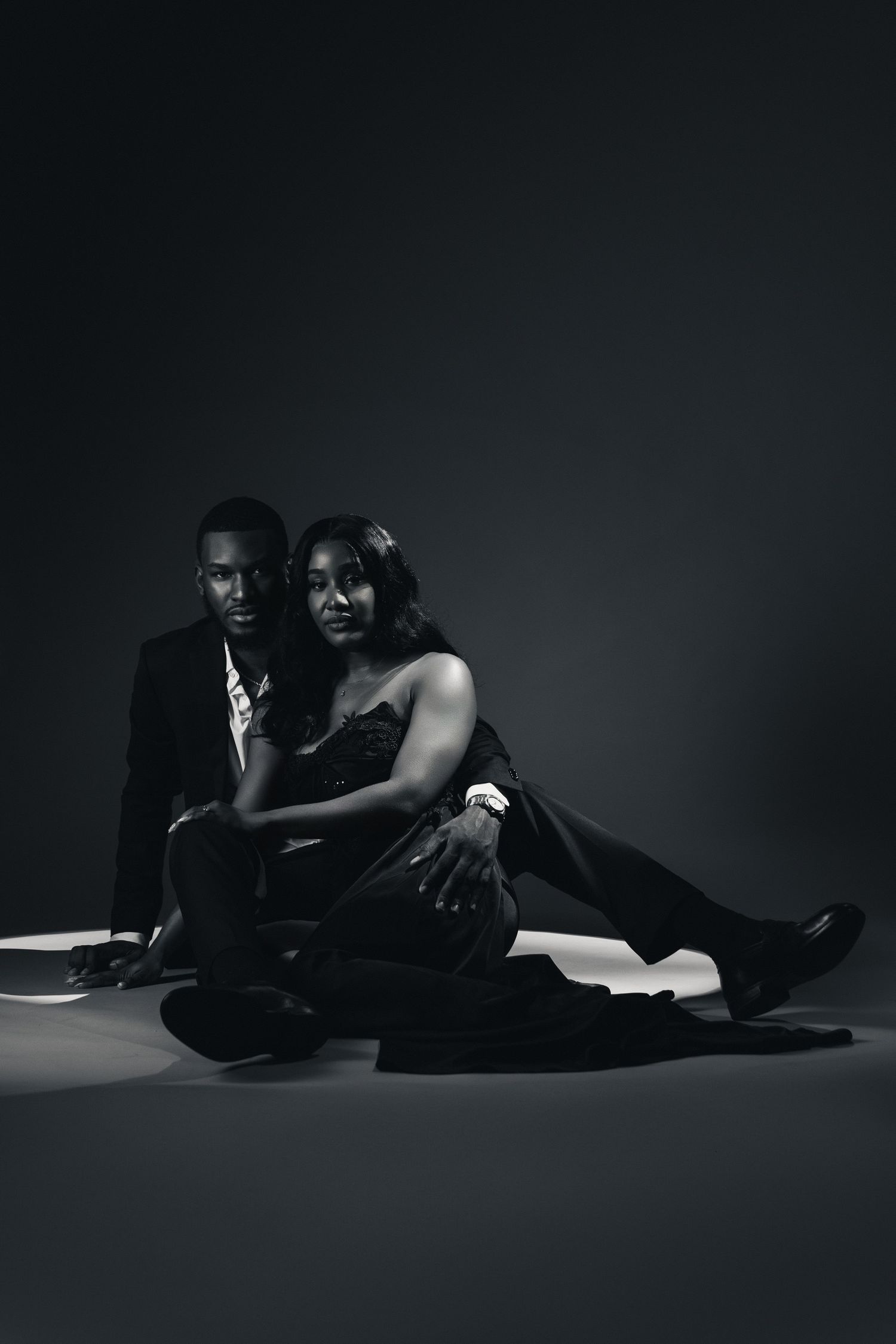 Elegant Black couple in formal attire sitting together on studio floor in dramatic black and white portrait photography.