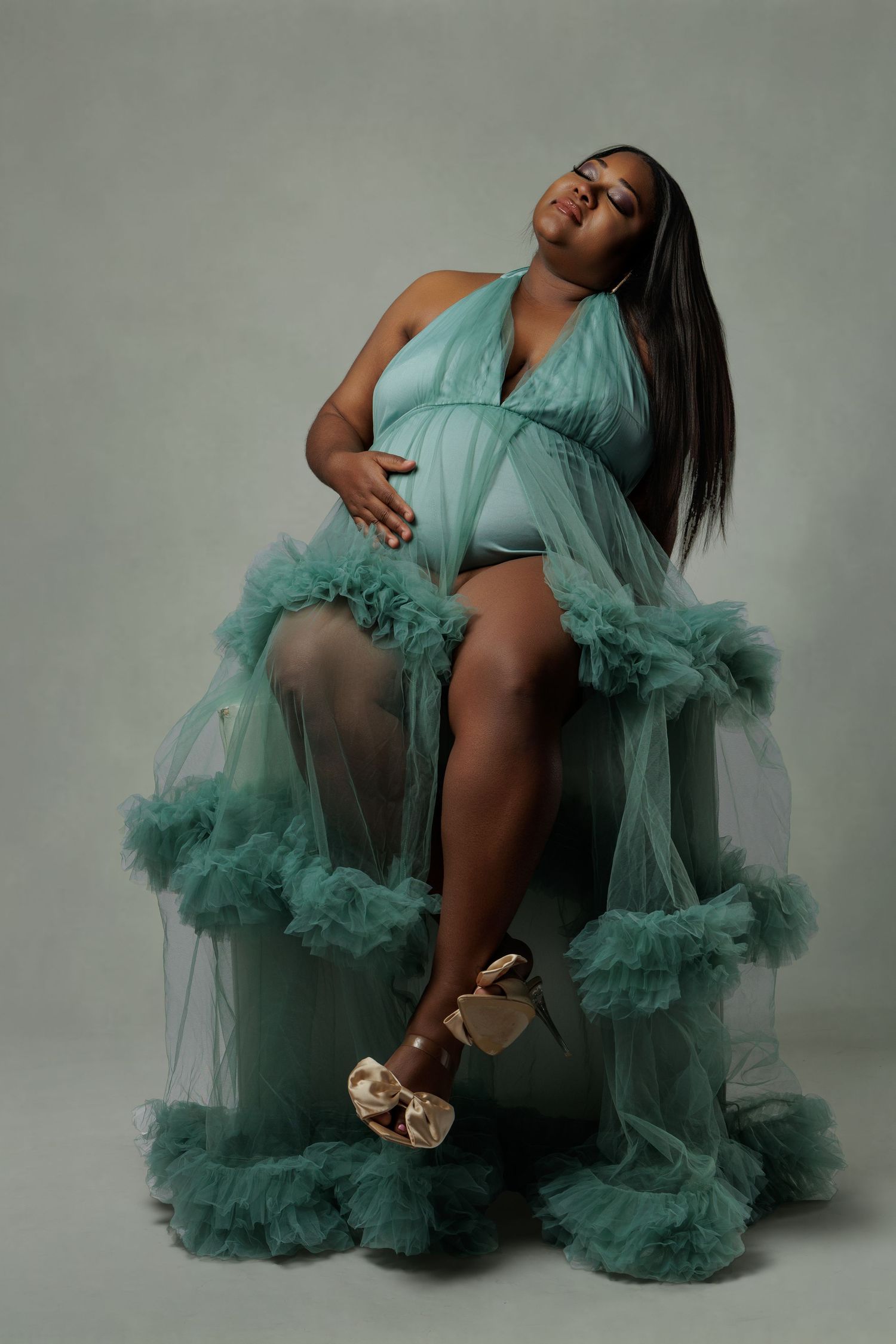 Pregnant woman in teal ruffle tulle maternity gown sitting gracefully, wearing gold heels in a studio portrait.