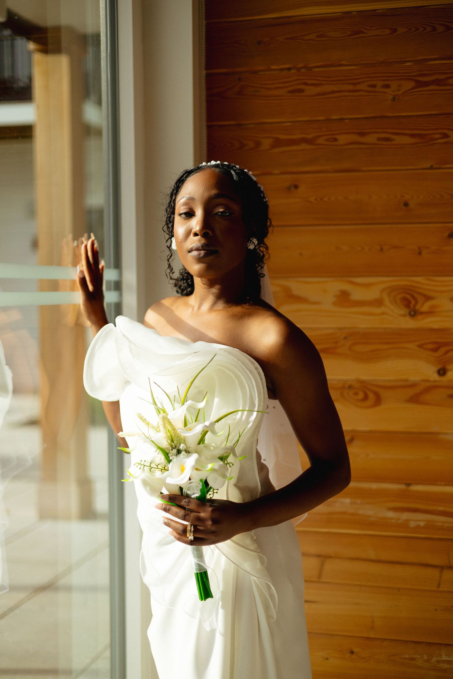 Elegant Black bride in a white one-shoulder gown holding a bouquet of white calla lilies by a window.