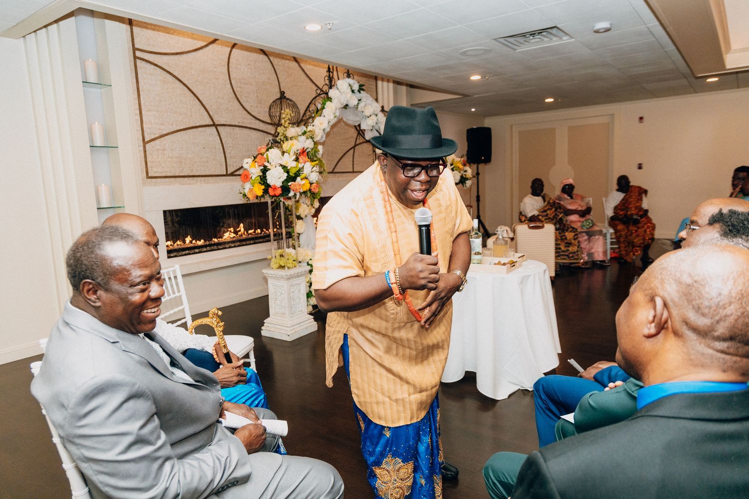 Man in orange shirt and hat speaks at an elegant indoor celebration event with guests seated nearby.