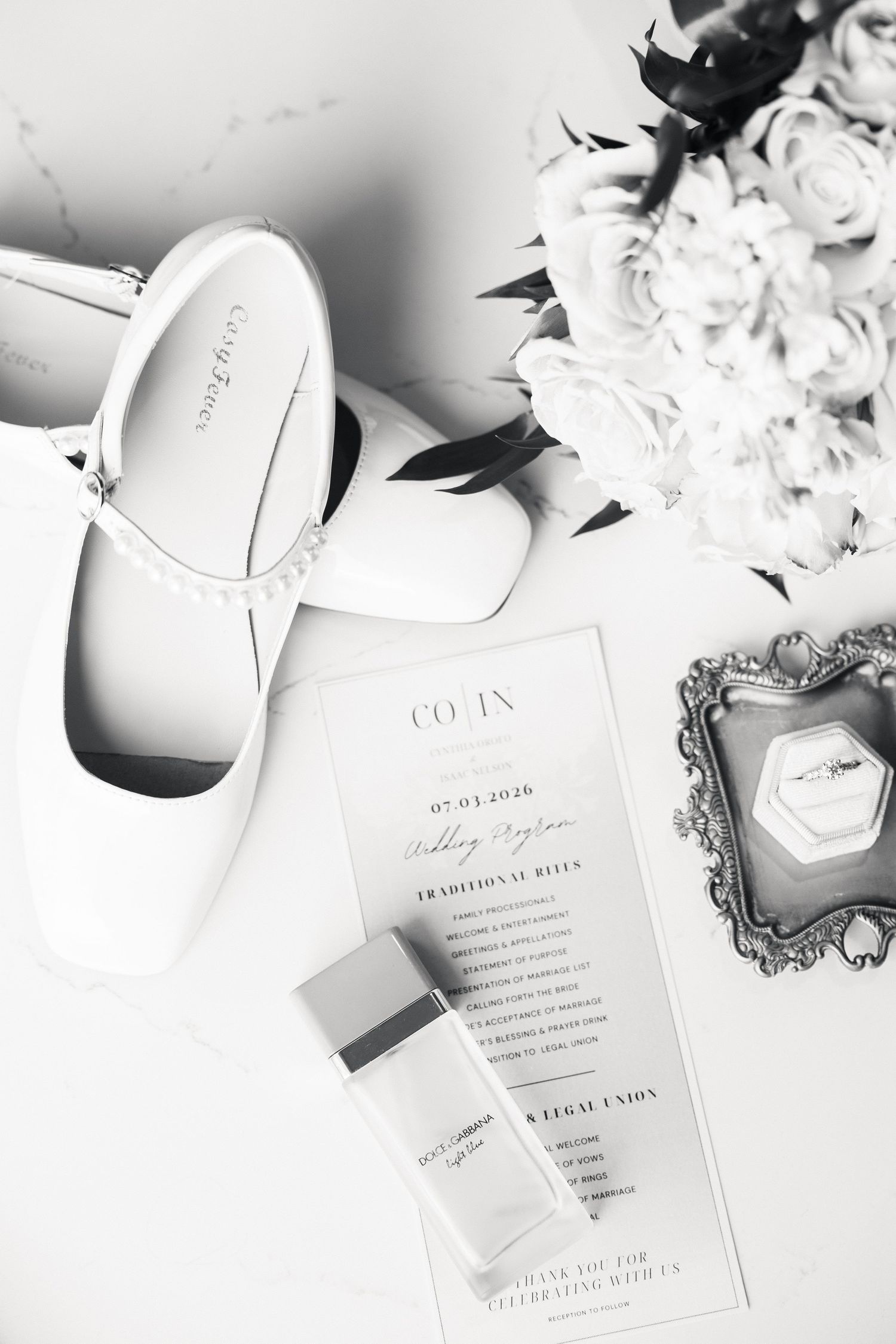 Black and white flat lay of bridal shoes, wedding program, perfume, and white roses bouquet on marble surface.