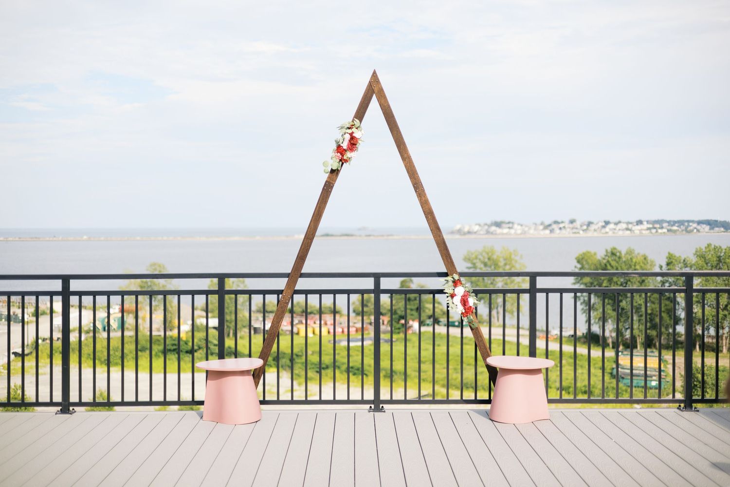 Wooden triangular wedding arch with floral accents on a waterfront deck overlooking a scenic bay.