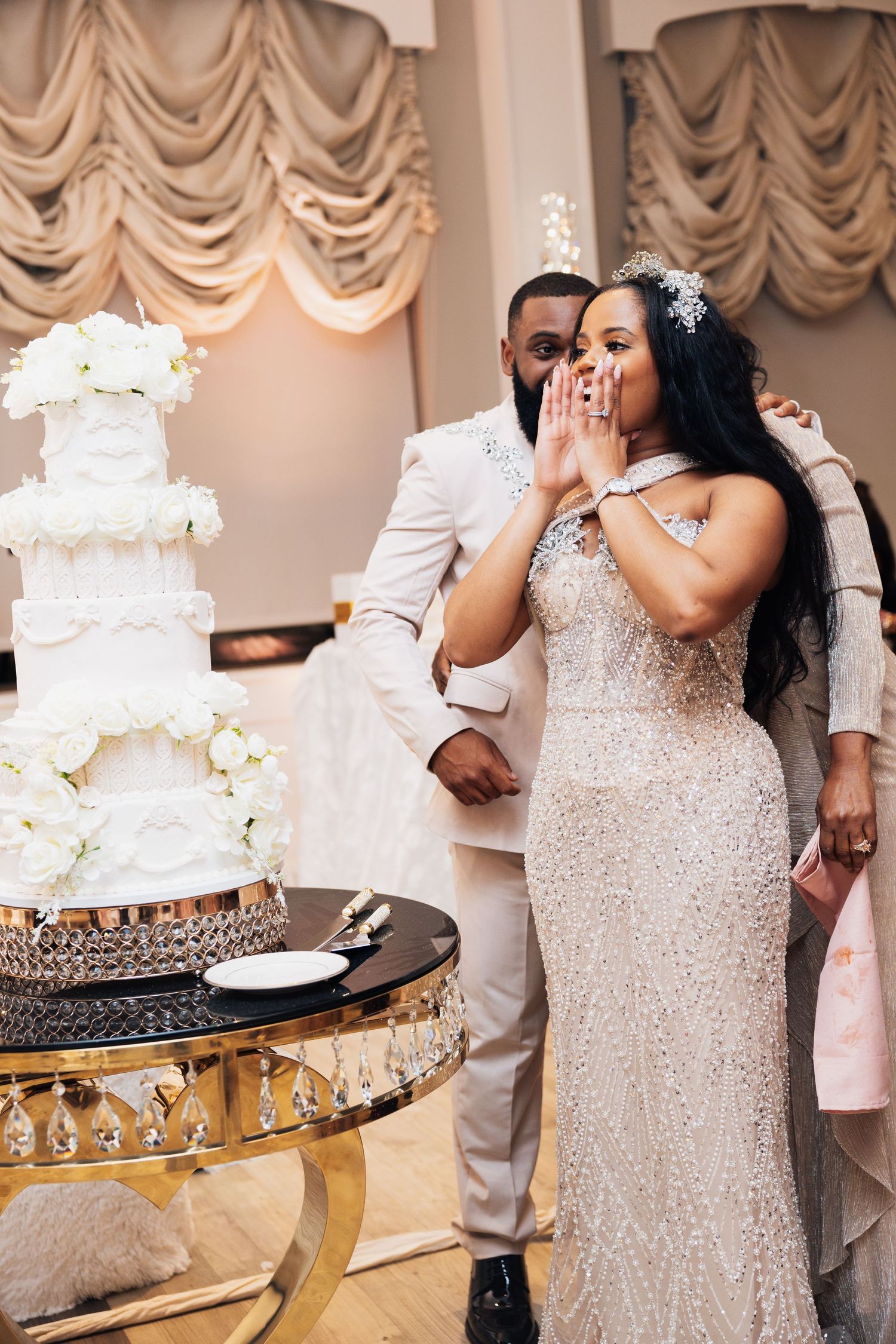 Bride and groom in elegant attire stand beside a tall white floral wedding cake at a luxurious reception venue.