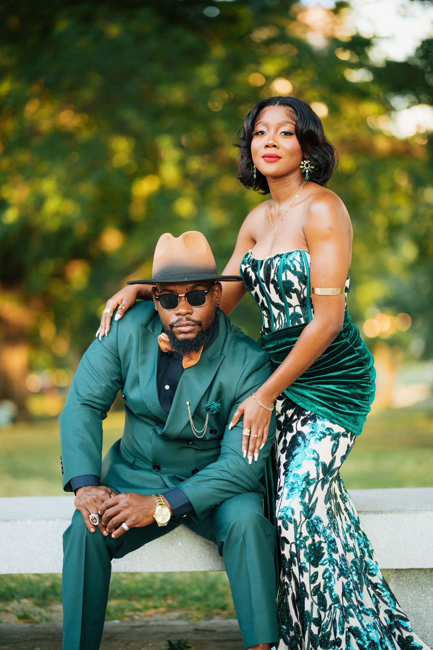 Elegant couple in matching green outfits posing in a park, man in suit and hat, woman in floral gown.