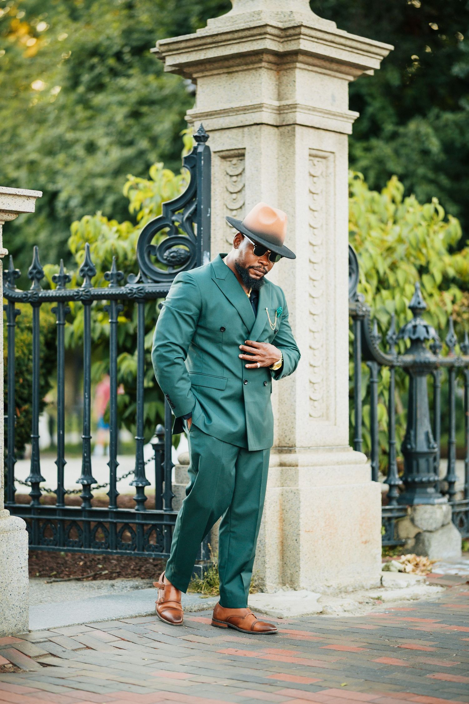 Stylish man in green suit, brown hat, sunglasses, and tan loafers posing by ornate iron gate outdoors.