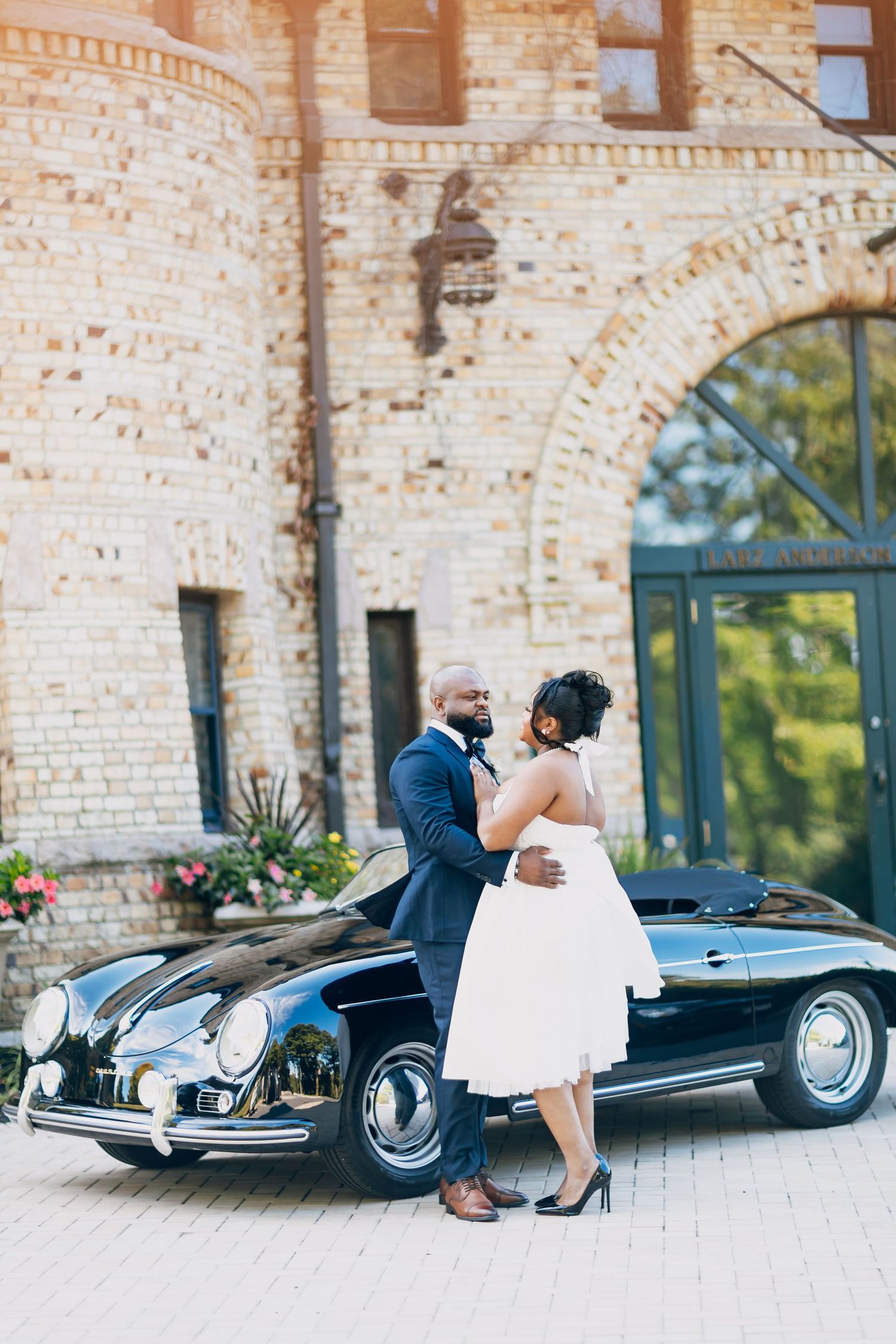 African American bride and groom embracing beside a vintage black Porsche at a brick wedding venue.