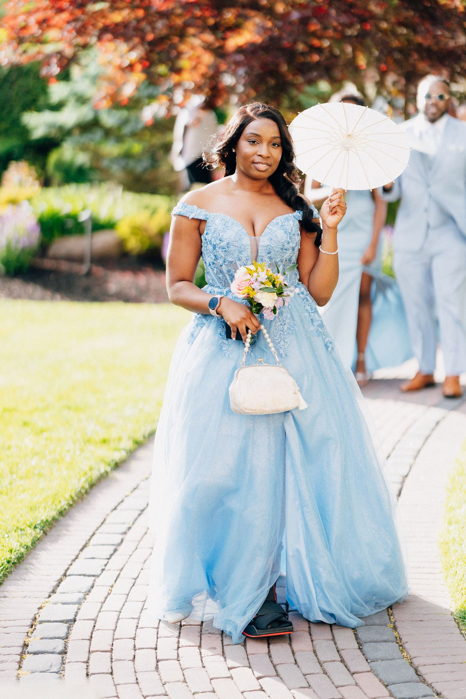 Bridesmaid in a blue off-shoulder gown holds a parasol and bouquet at an outdoor wedding.