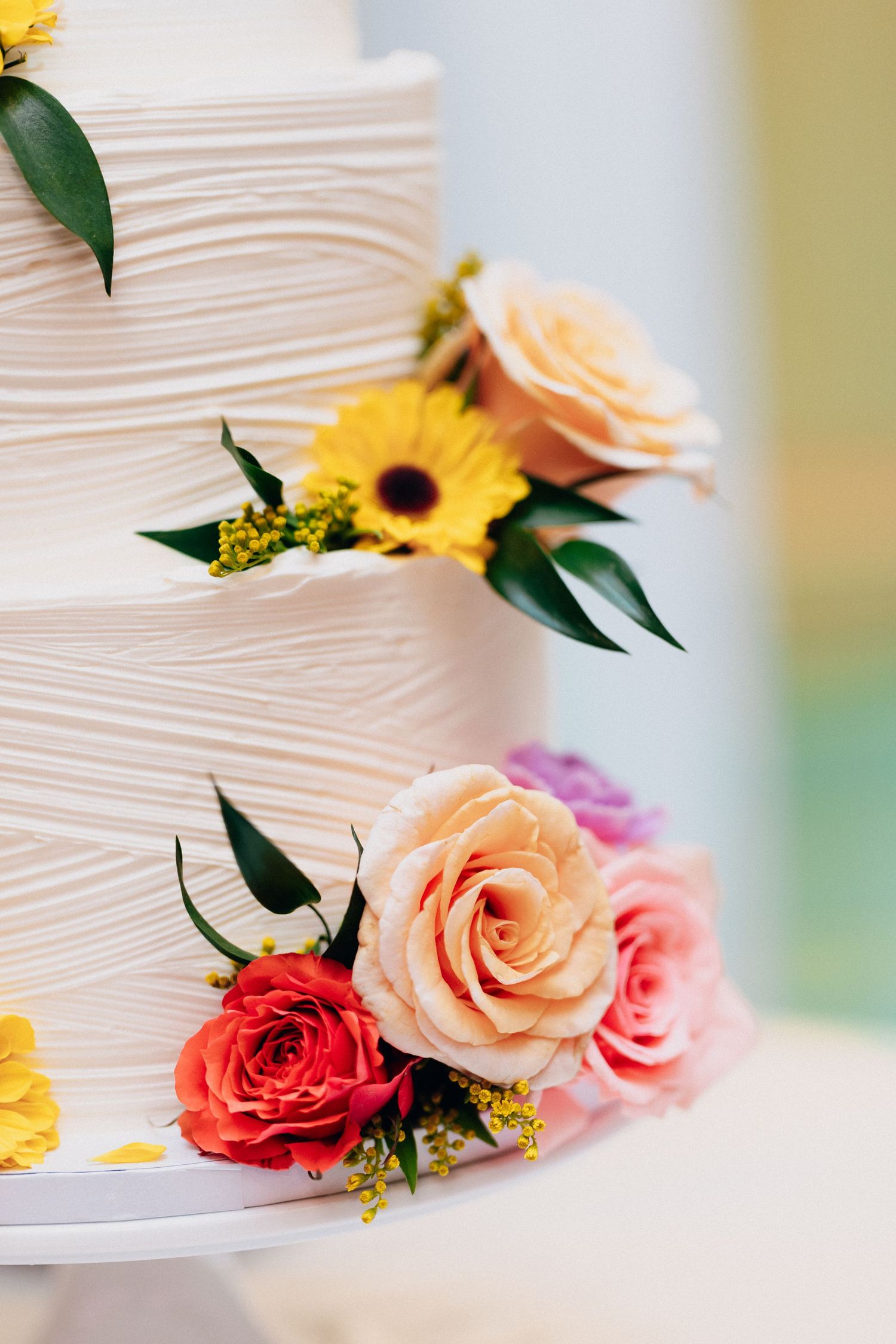 Elegant three-tier white wedding cake decorated with colorful roses, sunflowers, and greenery floral arrangements.