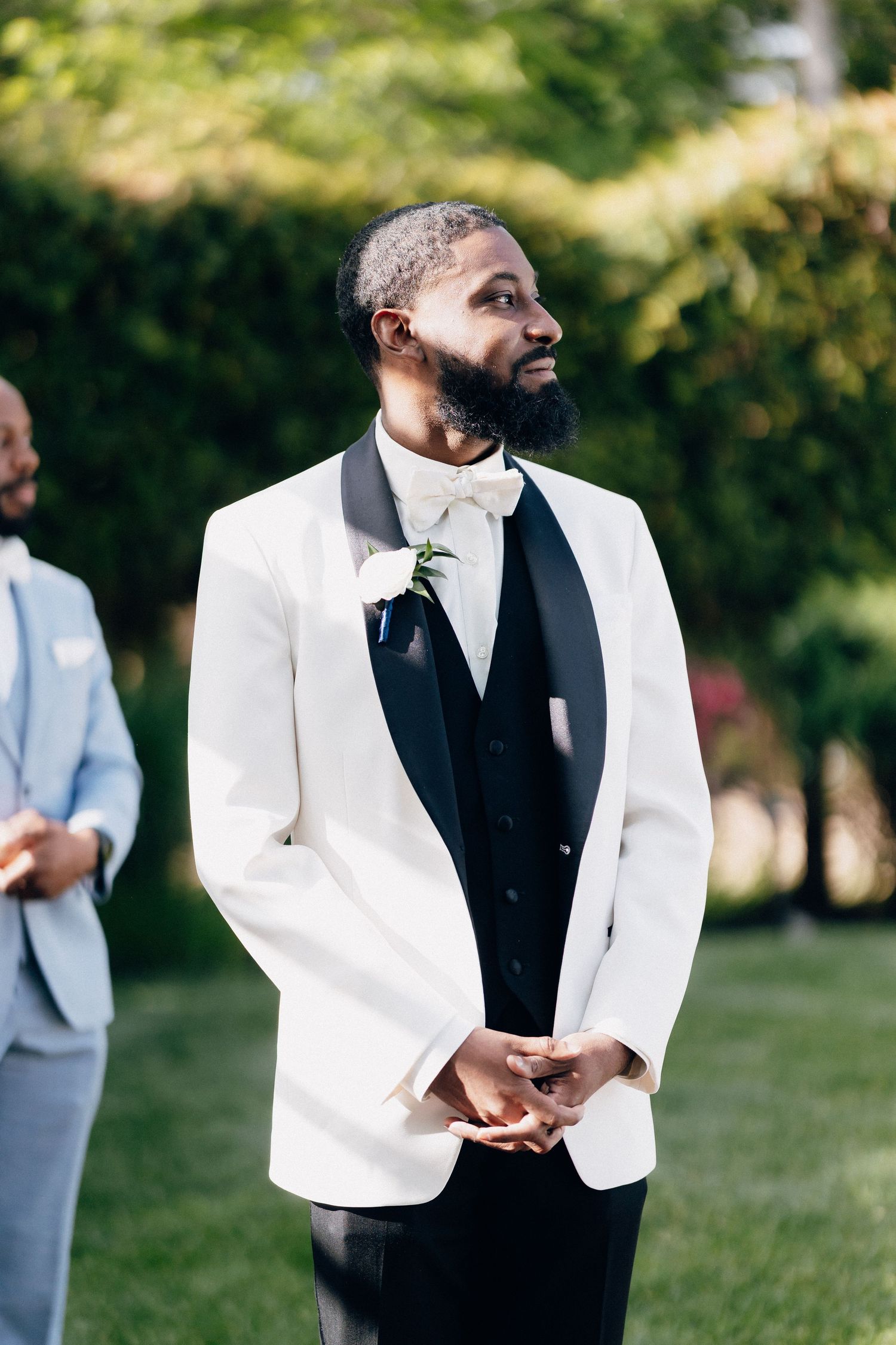 Groom in white tuxedo jacket with bow tie and boutonniere stands outdoors at wedding ceremony.