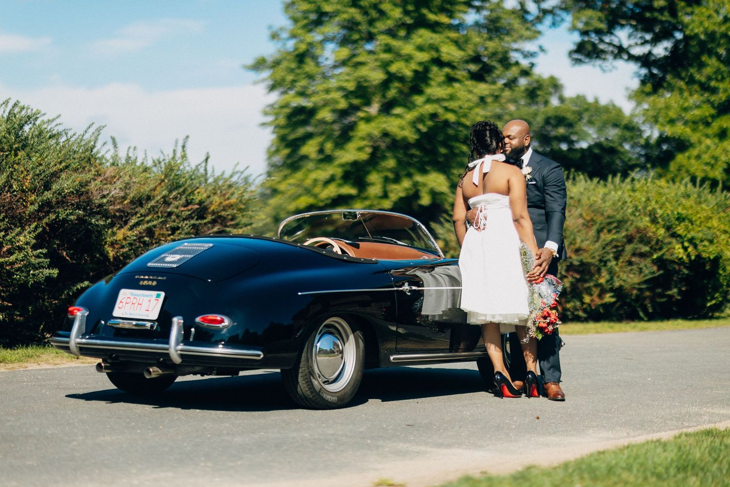 Bride and groom stand together behind a classic black convertible Porsche on a sunny day.