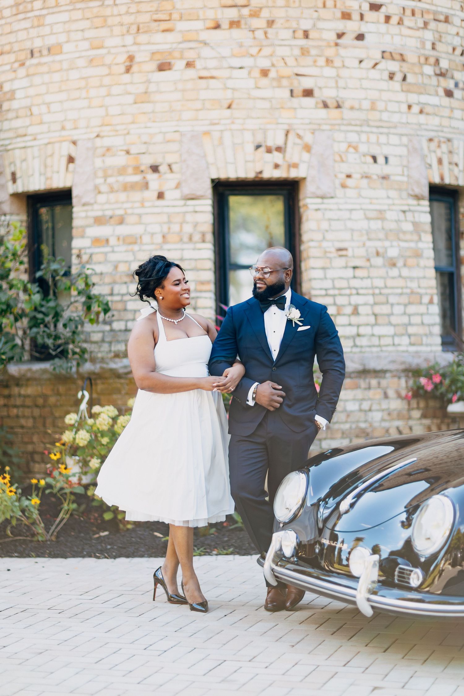 Black couple in wedding attire smiling beside a vintage car near a brick building.