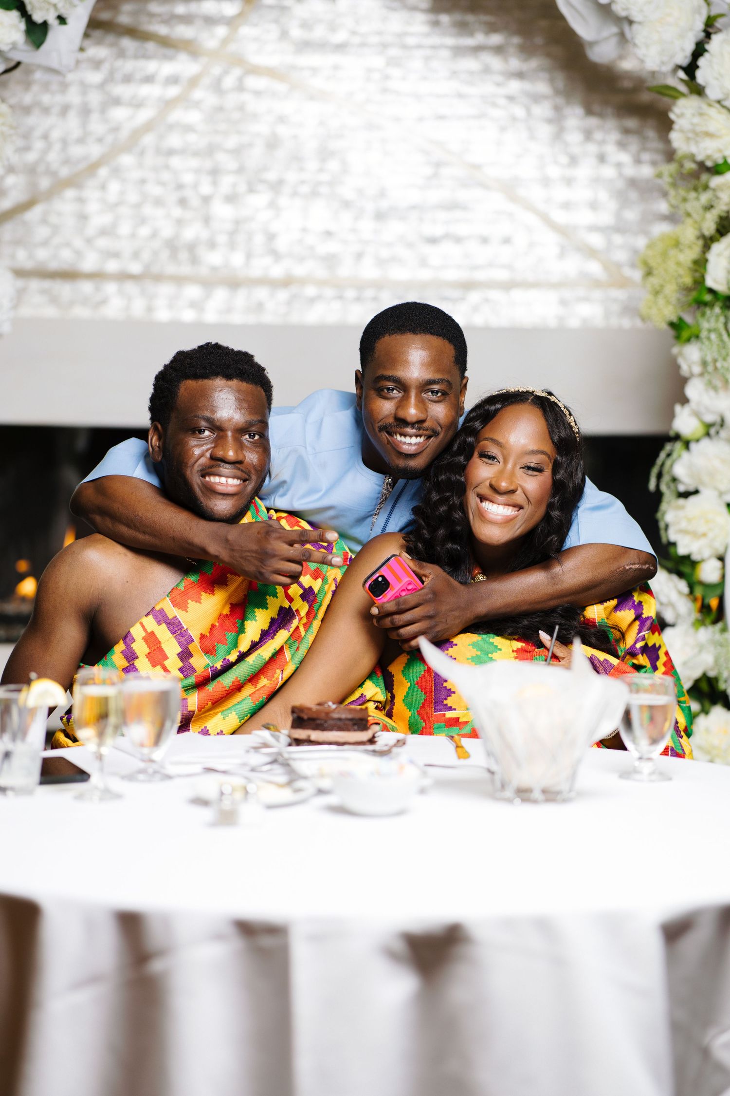 Two grooms in kente attire pose with friend at wedding reception table.