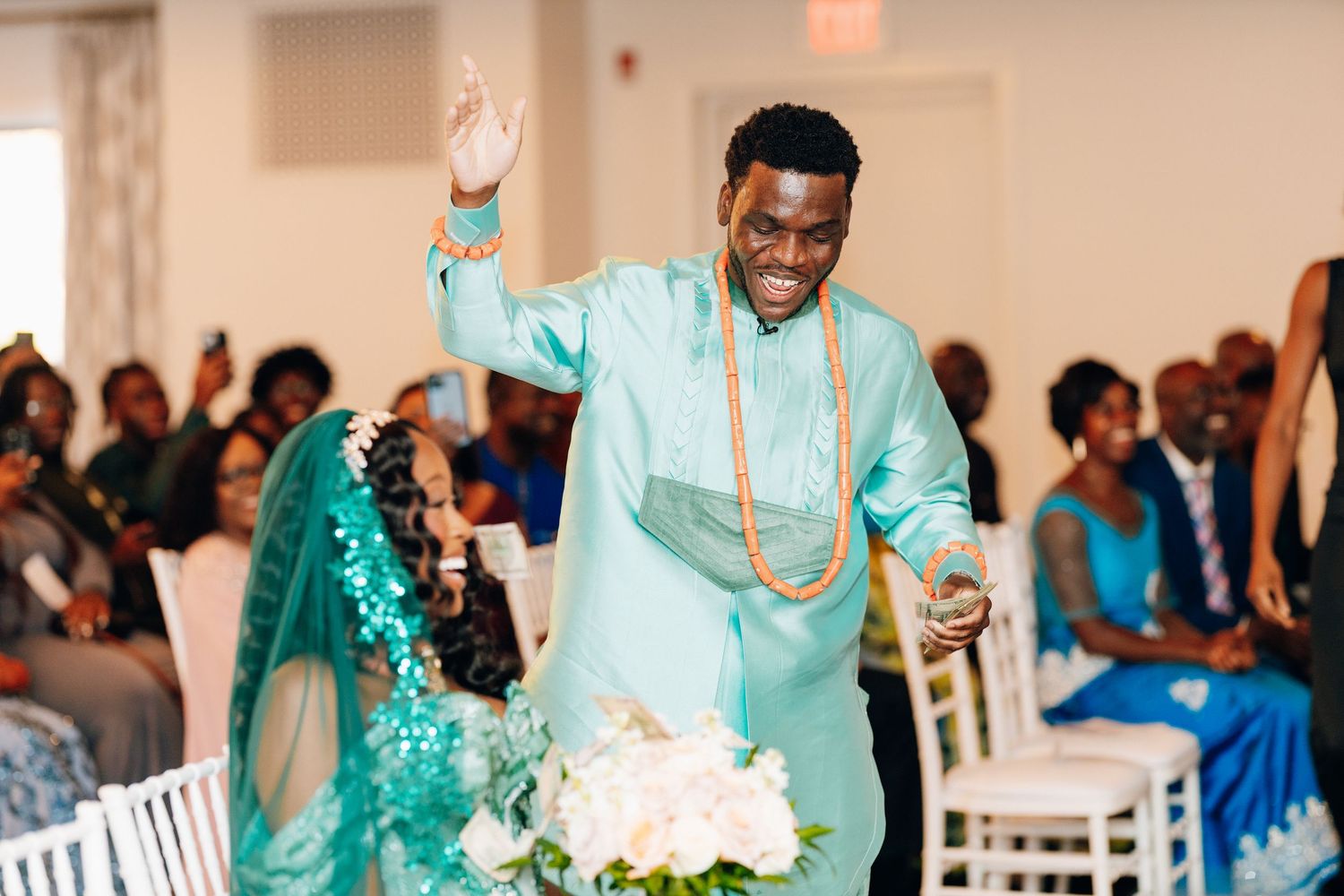 Groom in teal African attire raises hand joyfully while walking with bride at wedding.
