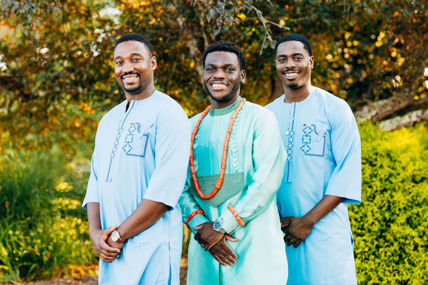 Three men in traditional African attire smile outdoors, with the center man wearing colorful beaded accessories.