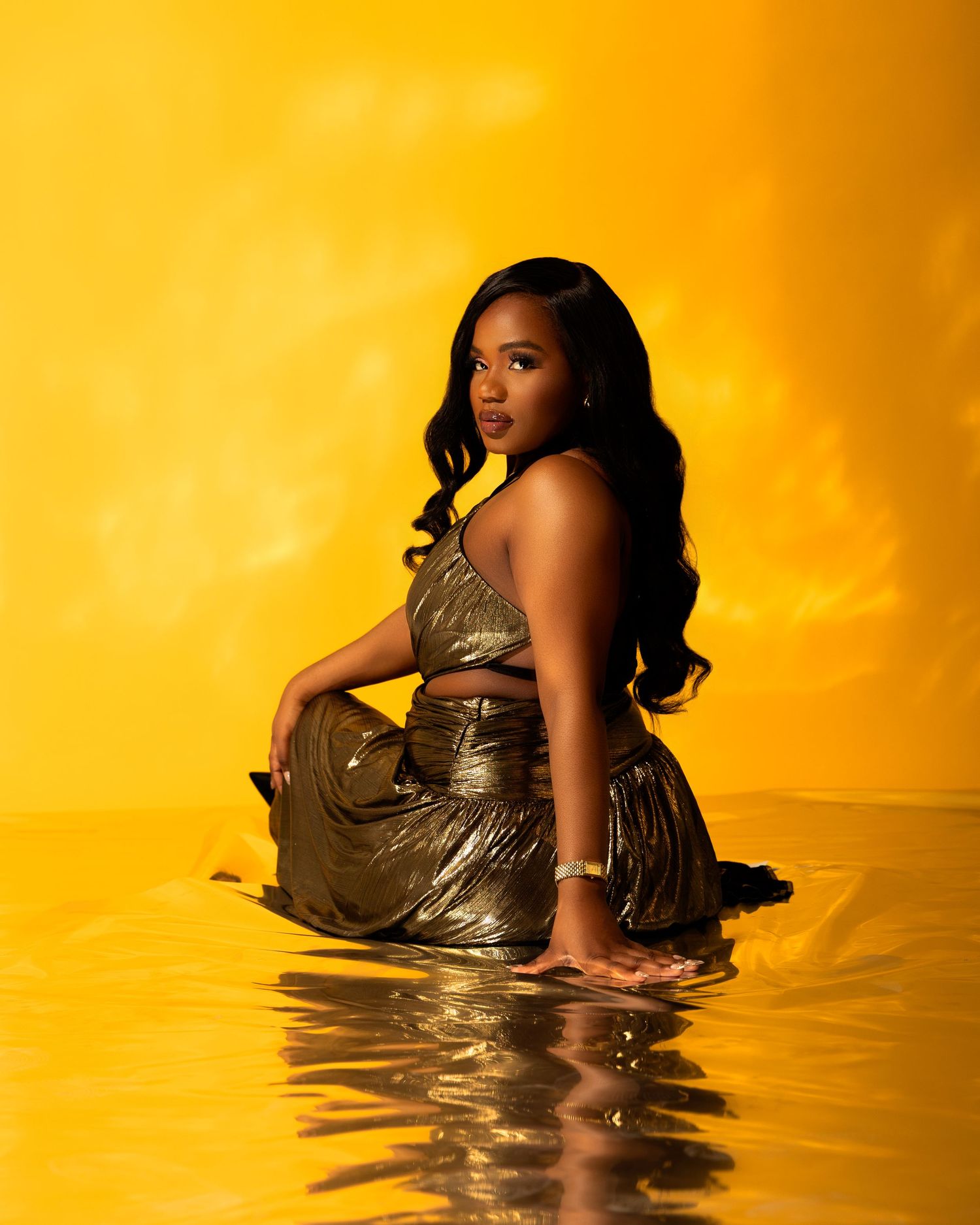 Woman in gold metallic outfit sitting gracefully in water against vibrant yellow background.